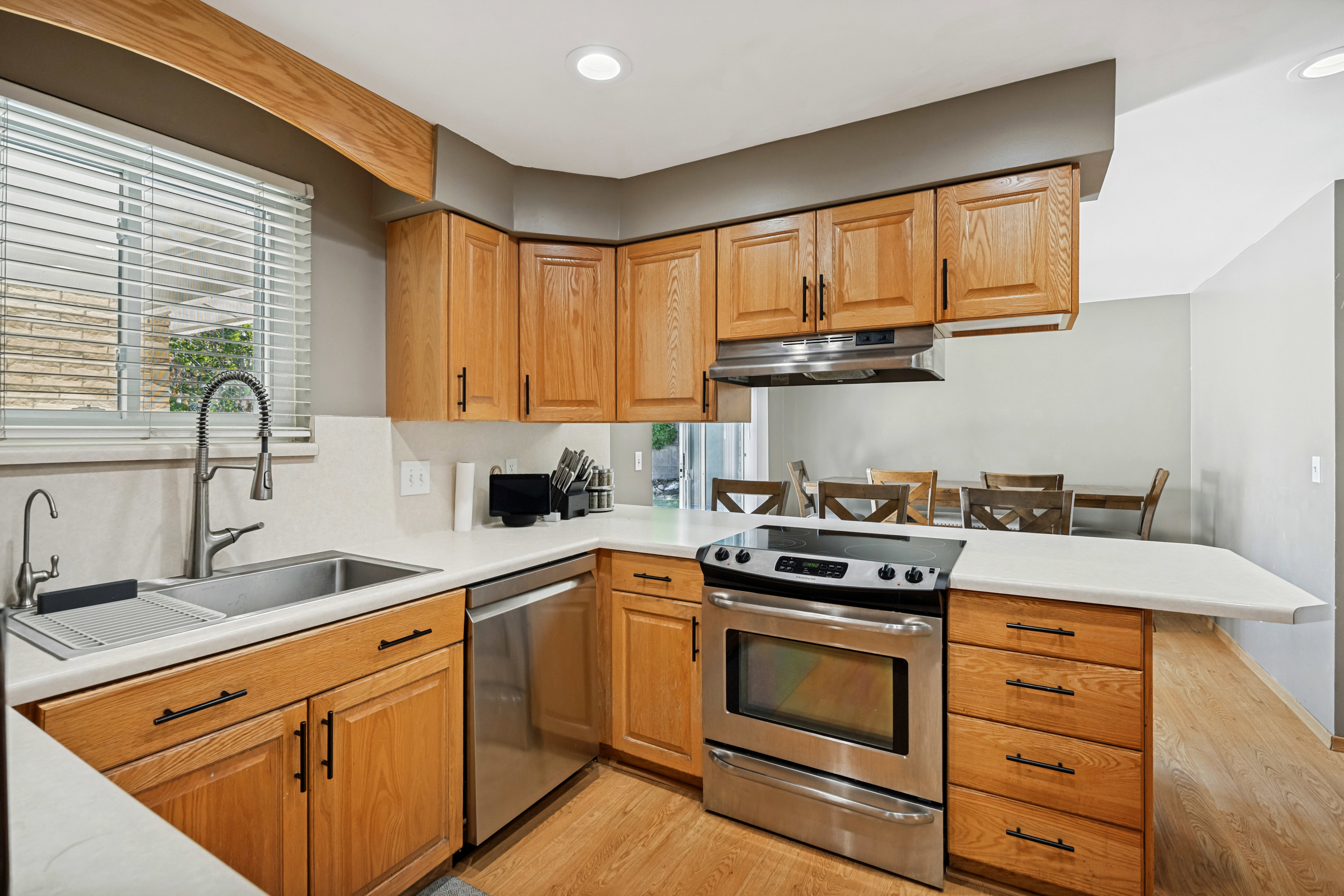 Kitchen featuring light wood cabinetry with dark hardware, stainless steel appliances including a range and dishwasher, a stainless steel sink with a gooseneck faucet, and light-toned countertops