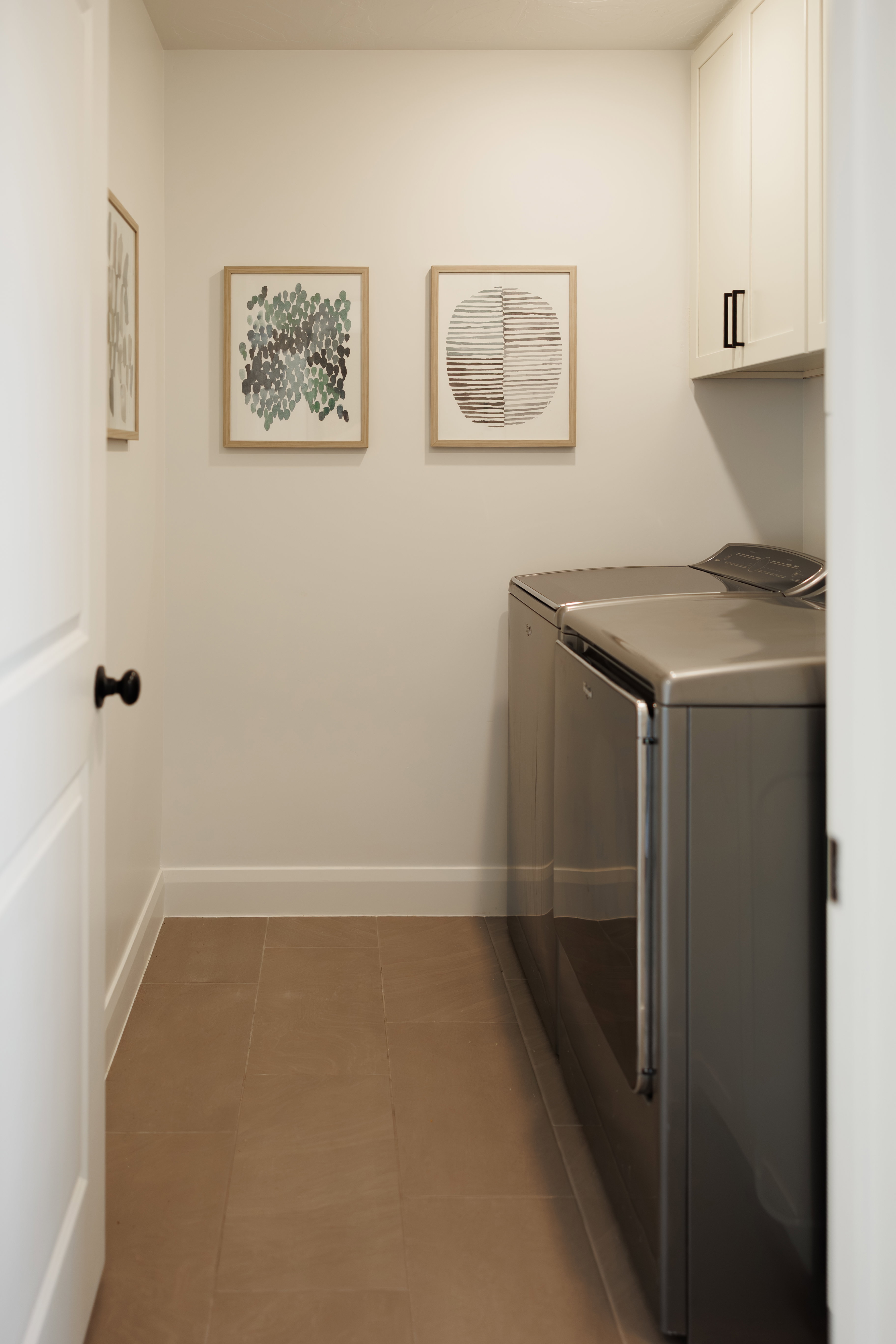 Laundry area featuring warm-toned tile flooring, built-in upper cabinetry with black hardware, and crisp white baseboards