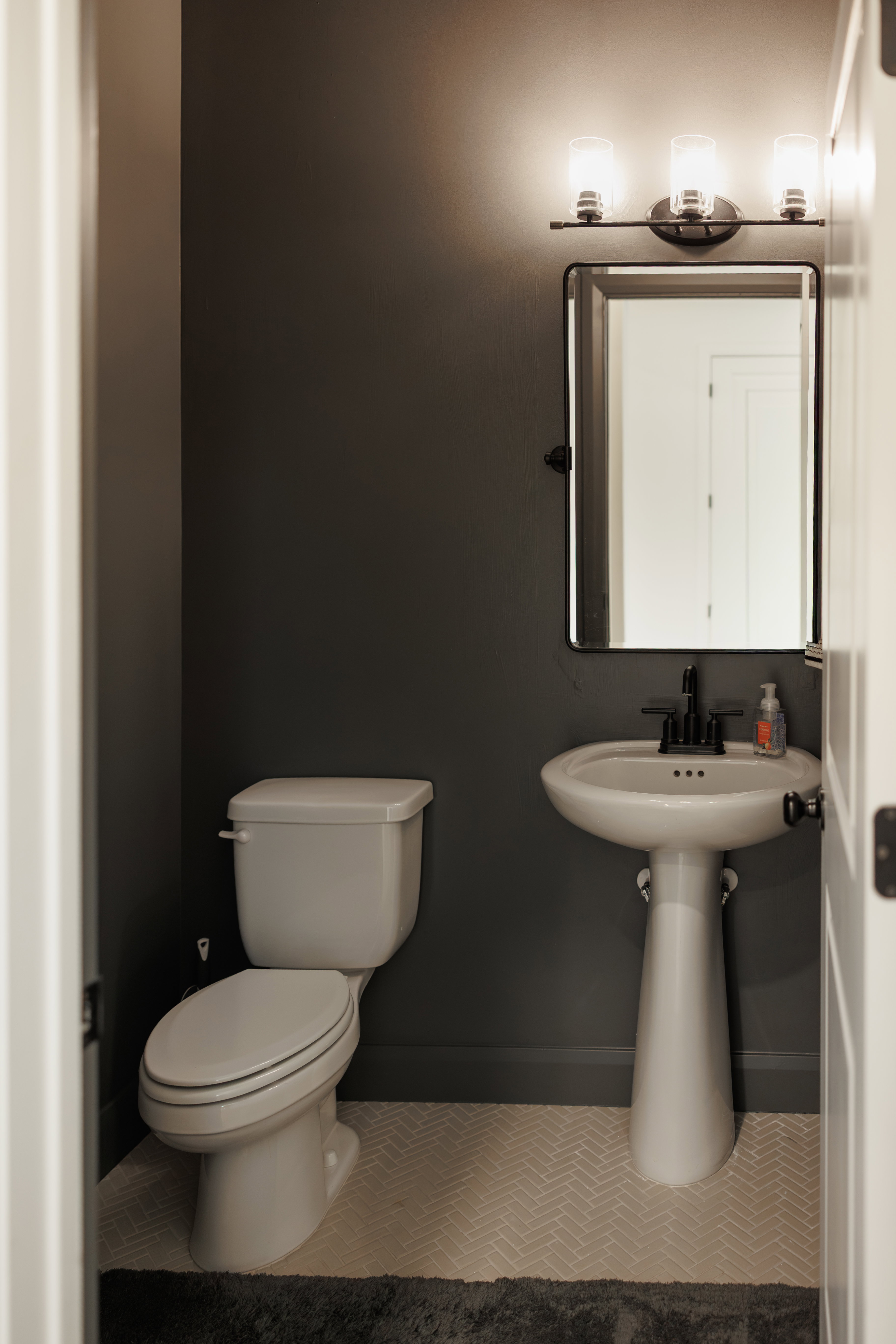 Compact lavatory featuring a white pedestal sink with a matte black faucet, a rectangular mirror, and a three-light vanity fixture