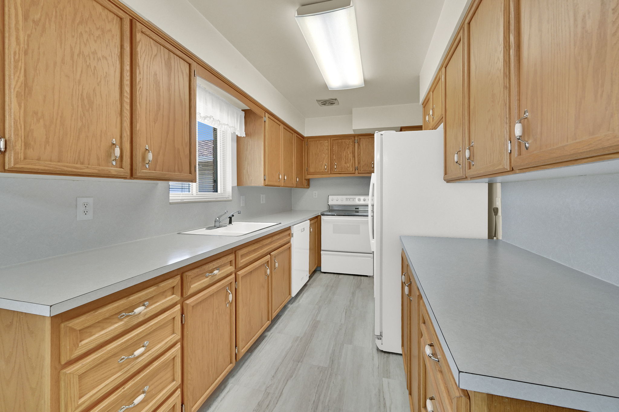 Galley kitchen featuring wood cabinetry, light countertops, wood-finish flooring, white appliances, and a window above the sink