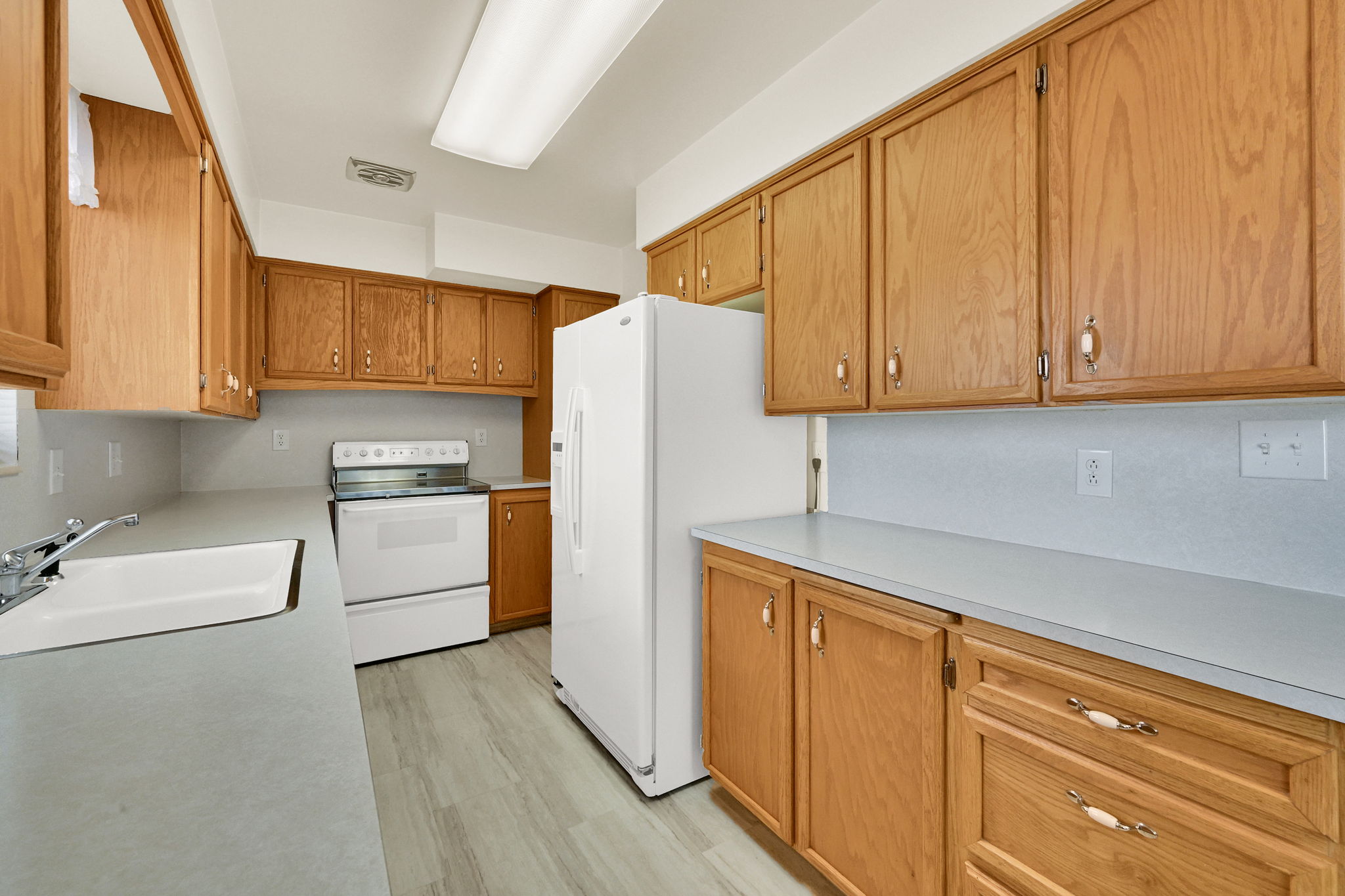 Galley kitchen featuring wood-finish cabinetry, light-toned countertops, and a white sink with chrome faucet