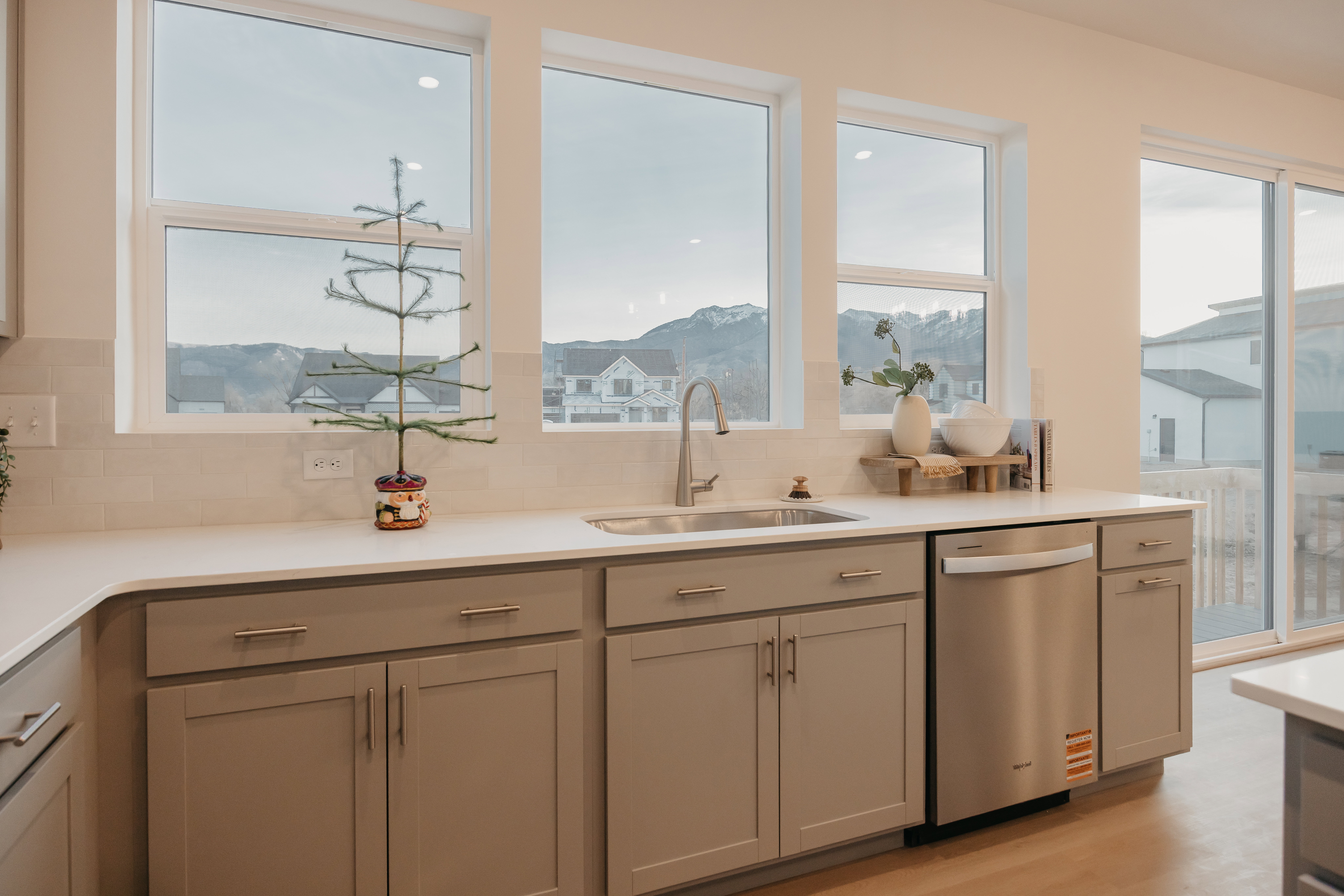 Kitchen featuring ample countertop space, grey cabinetry with brushed nickel hardware, an undermount stainless steel sink with a gooseneck faucet, a built-in stainless steel dishwasher, and large windows providing natural light