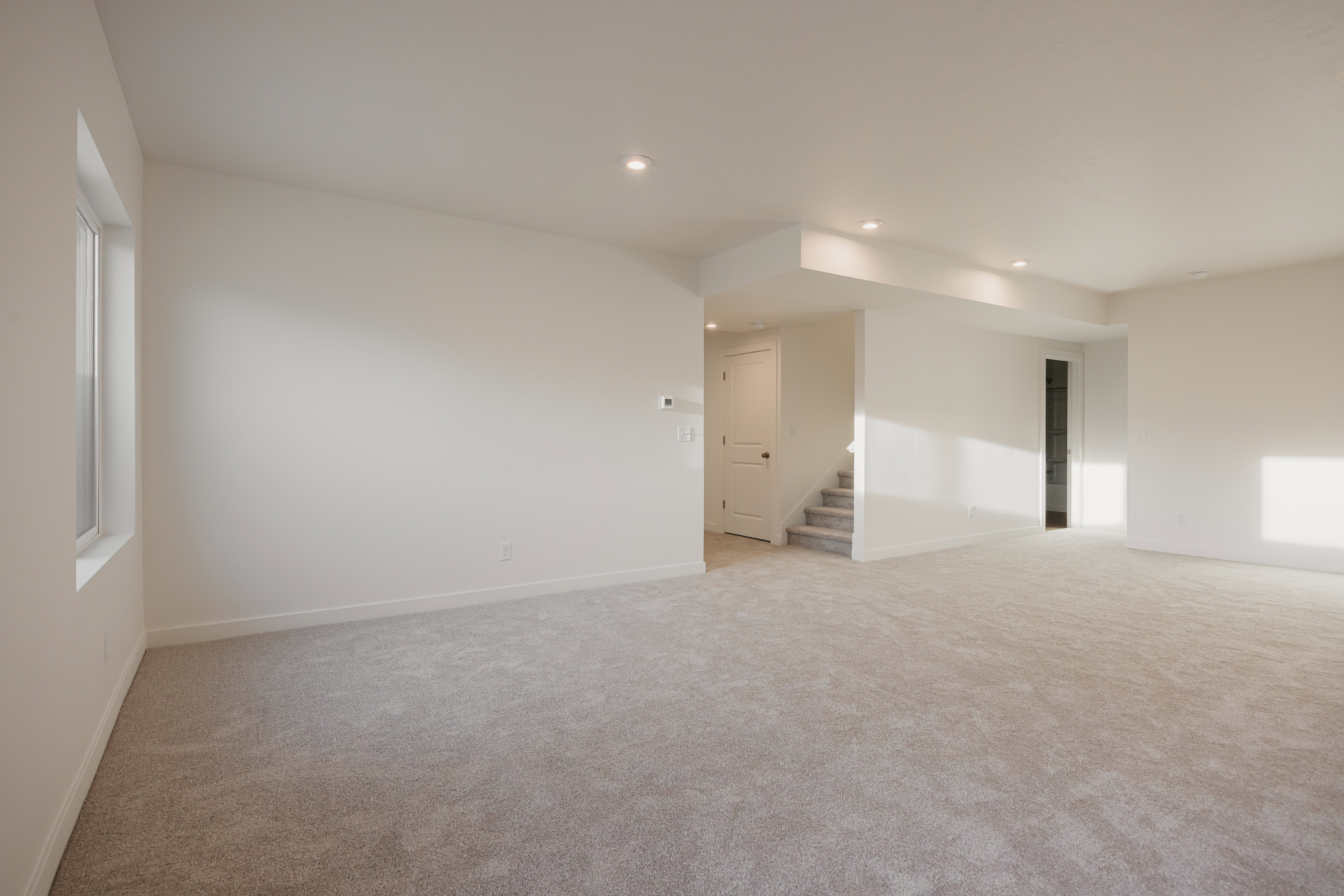 Expansive carpeted room featuring recessed lighting, a window with white trim, a staircase with carpeted treads, and a white paneled door