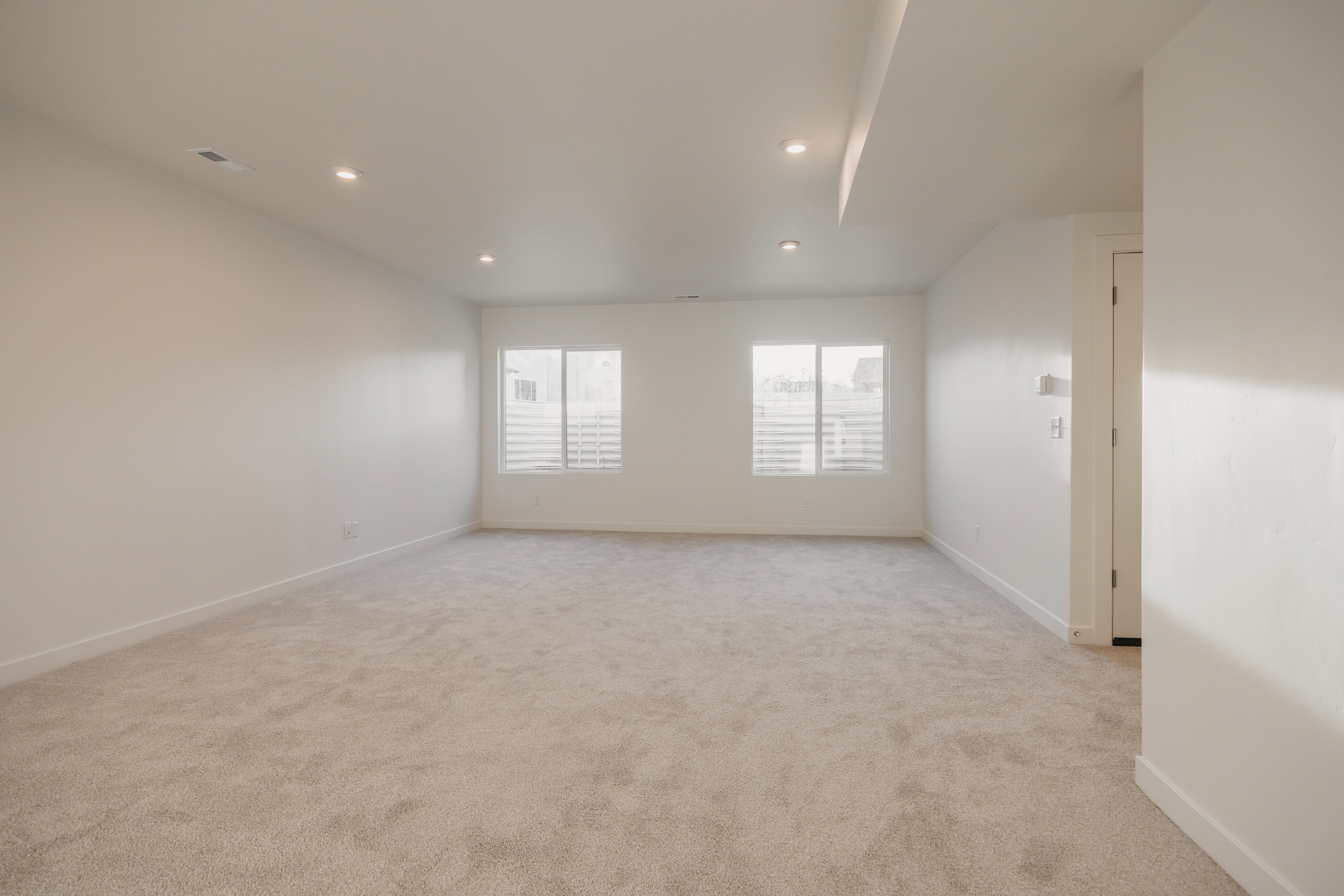 Spacious room featuring neutral carpeting, recessed ceiling lighting, two windows with horizontal blinds, and a white interior door