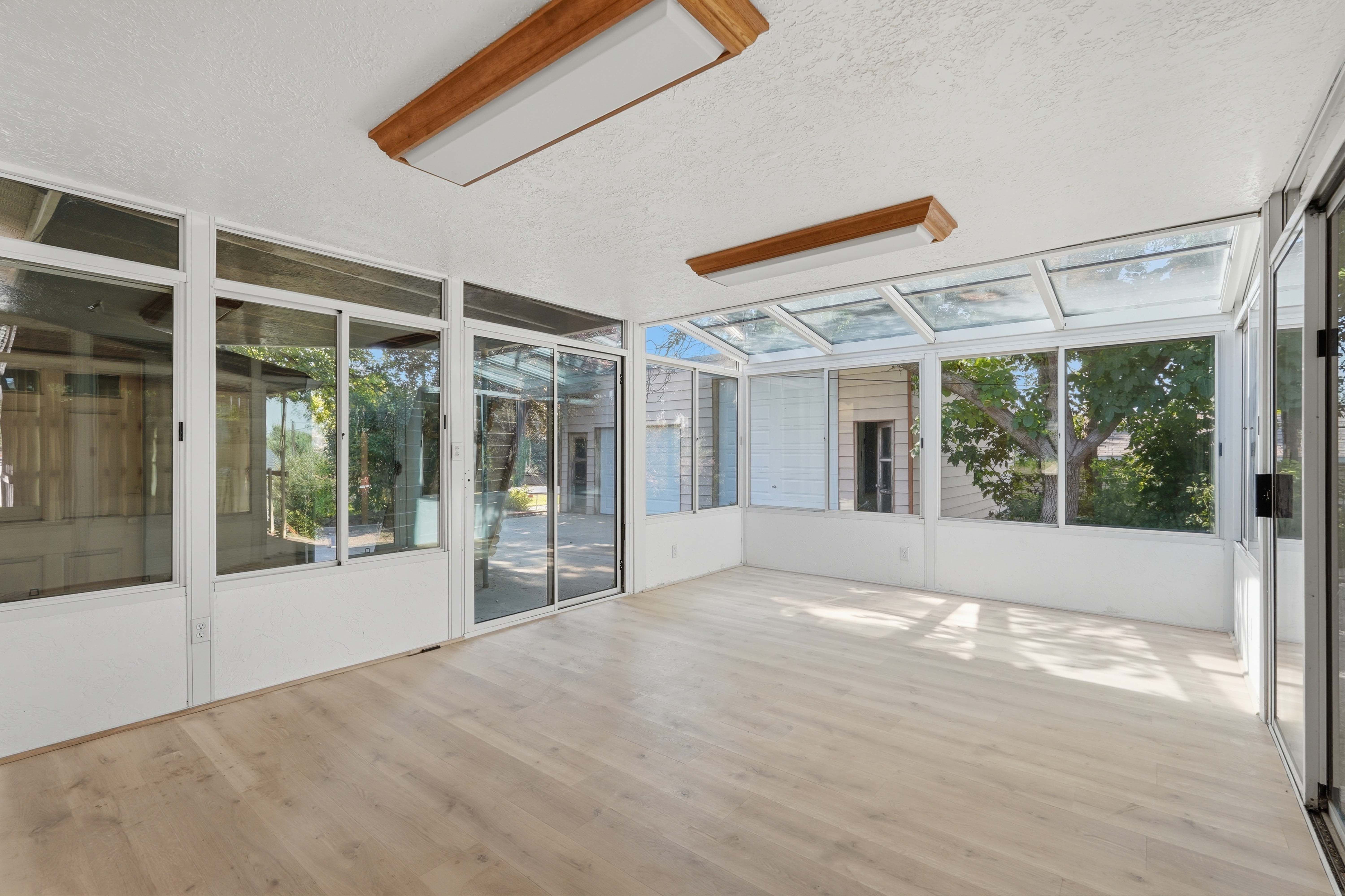 Sunroom featuring extensive glazing, a vaulted glass ceiling, wood-finish flooring, two rectangular fluorescent light fixtures, and sliding glass doors