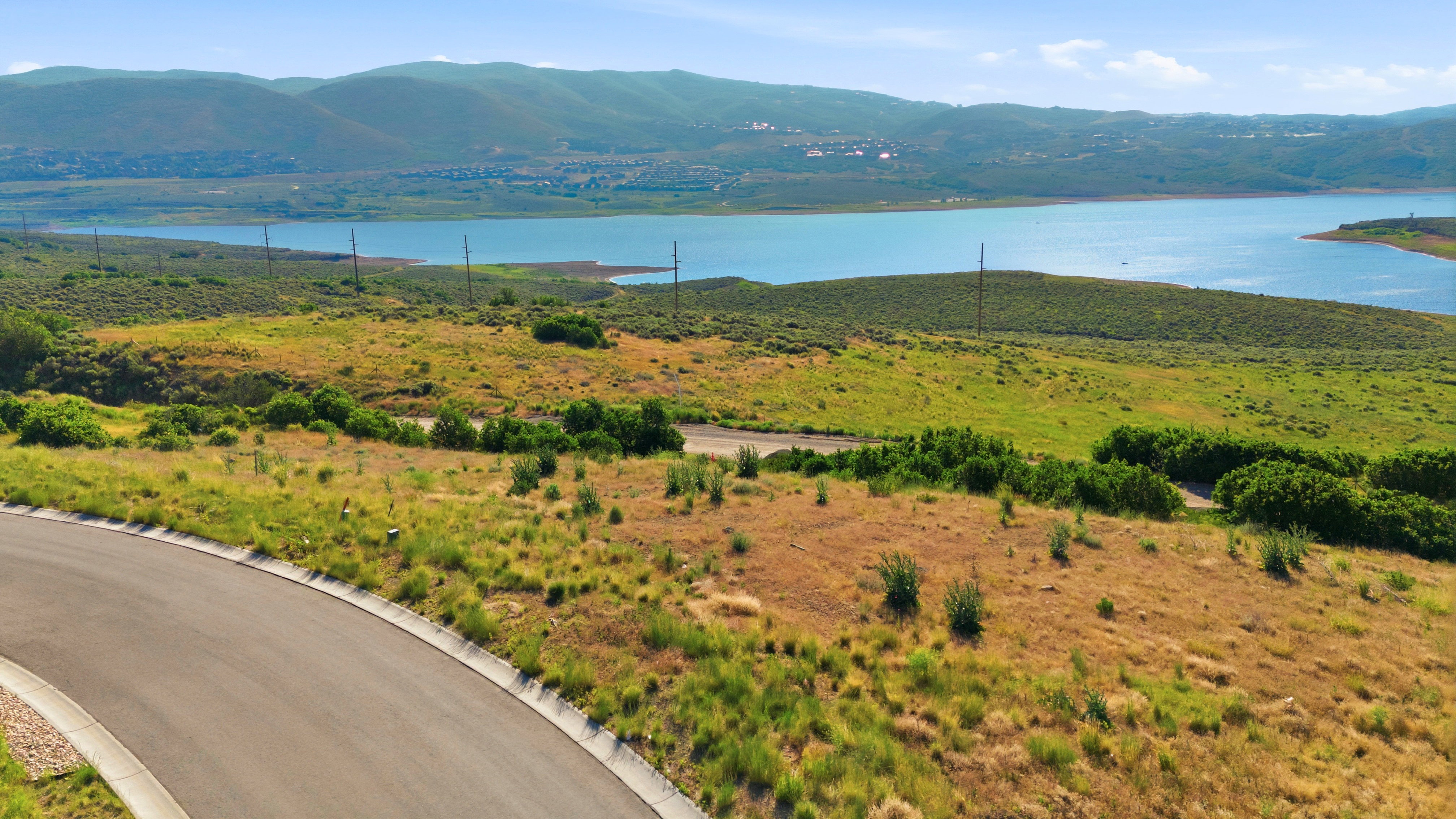 Paved road bordering an expanse of natural terrain, featuring varied vegetation, a prominent body of water, and distant mountain ranges under a clear sky