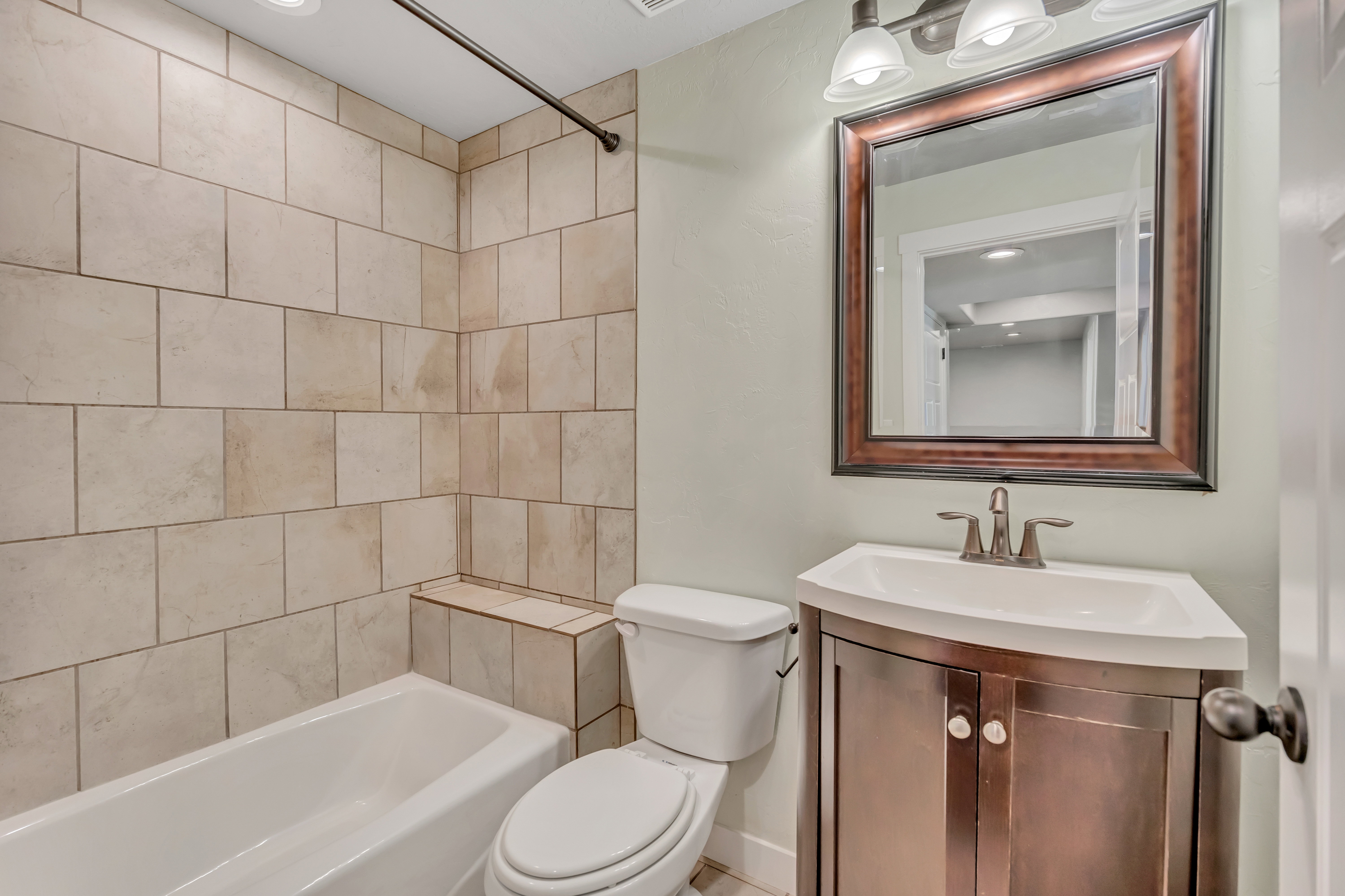 Full bathroom featuring a tiled shower surround with a built-in bench, a white porcelain bathtub, a curved vanity with a white countertop and integrated sink, a framed mirror, and a wall-mounted light fixture