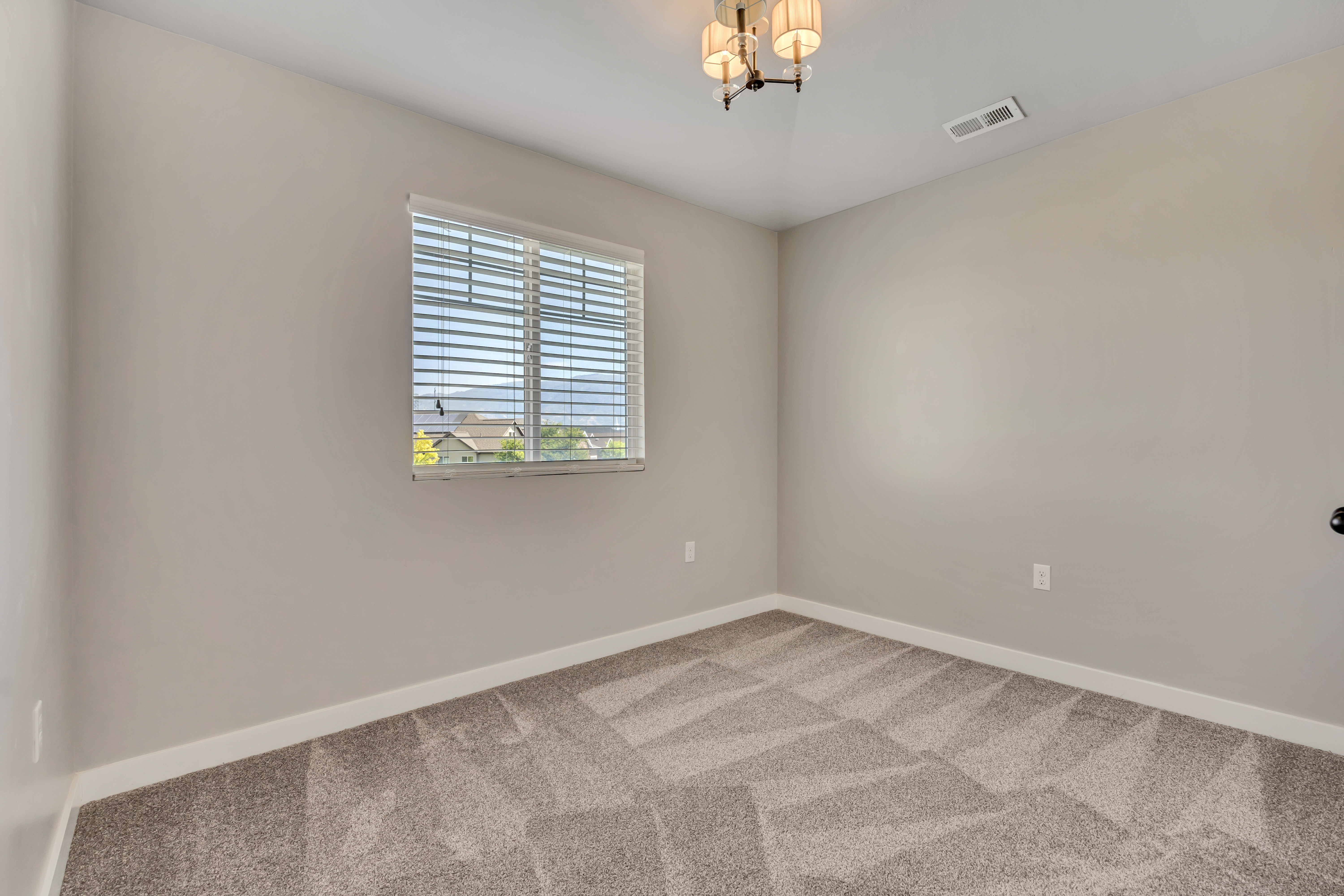 Neutral-toned room featuring a single window with horizontal blinds, overhead chandelier lighting, and geometric-patterned carpeting