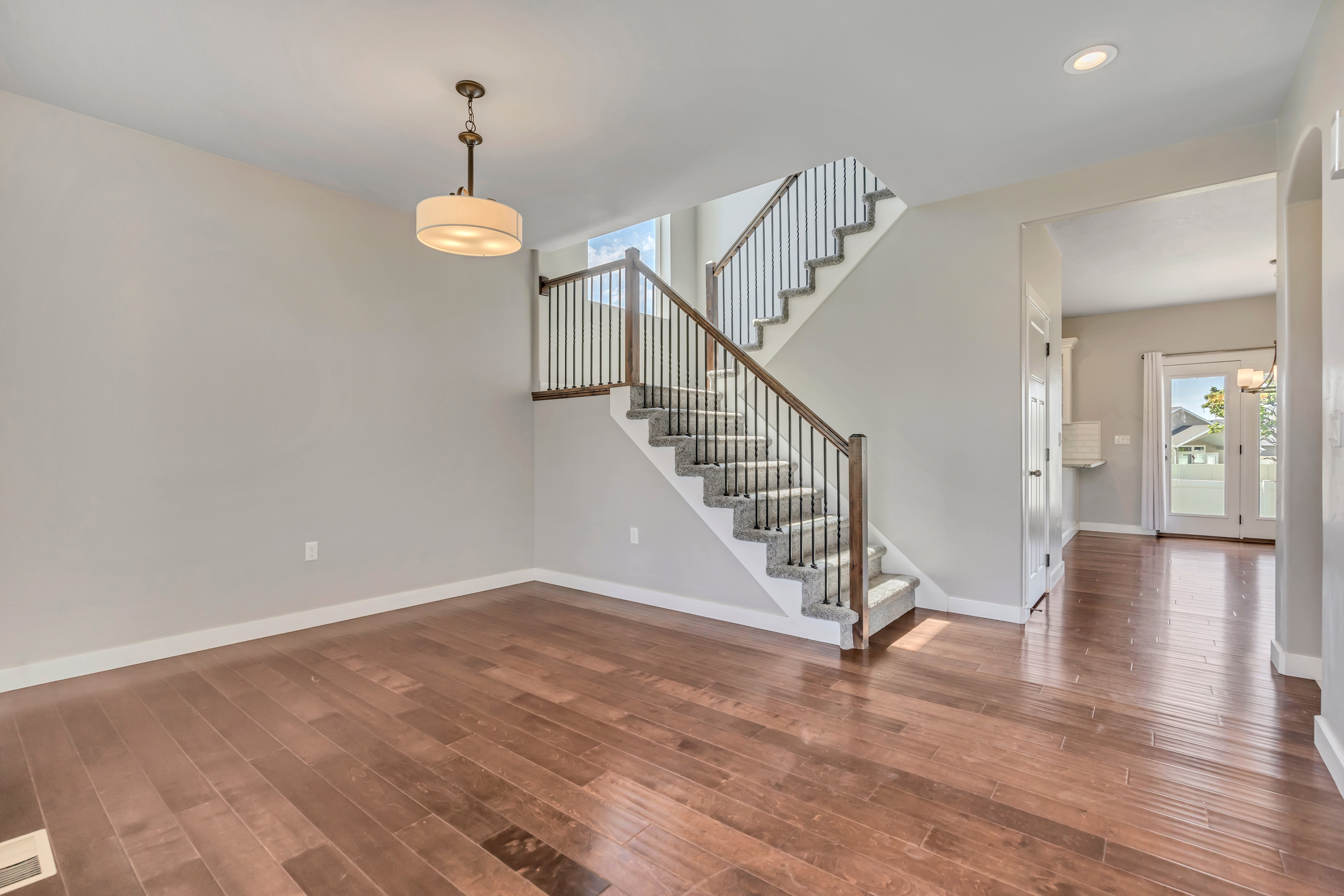 Hardwood-finish flooring extending through the entry, featuring a carpeted staircase with dark wood handrails and black metal balusters, a drum pendant light, and a white trim baseboard