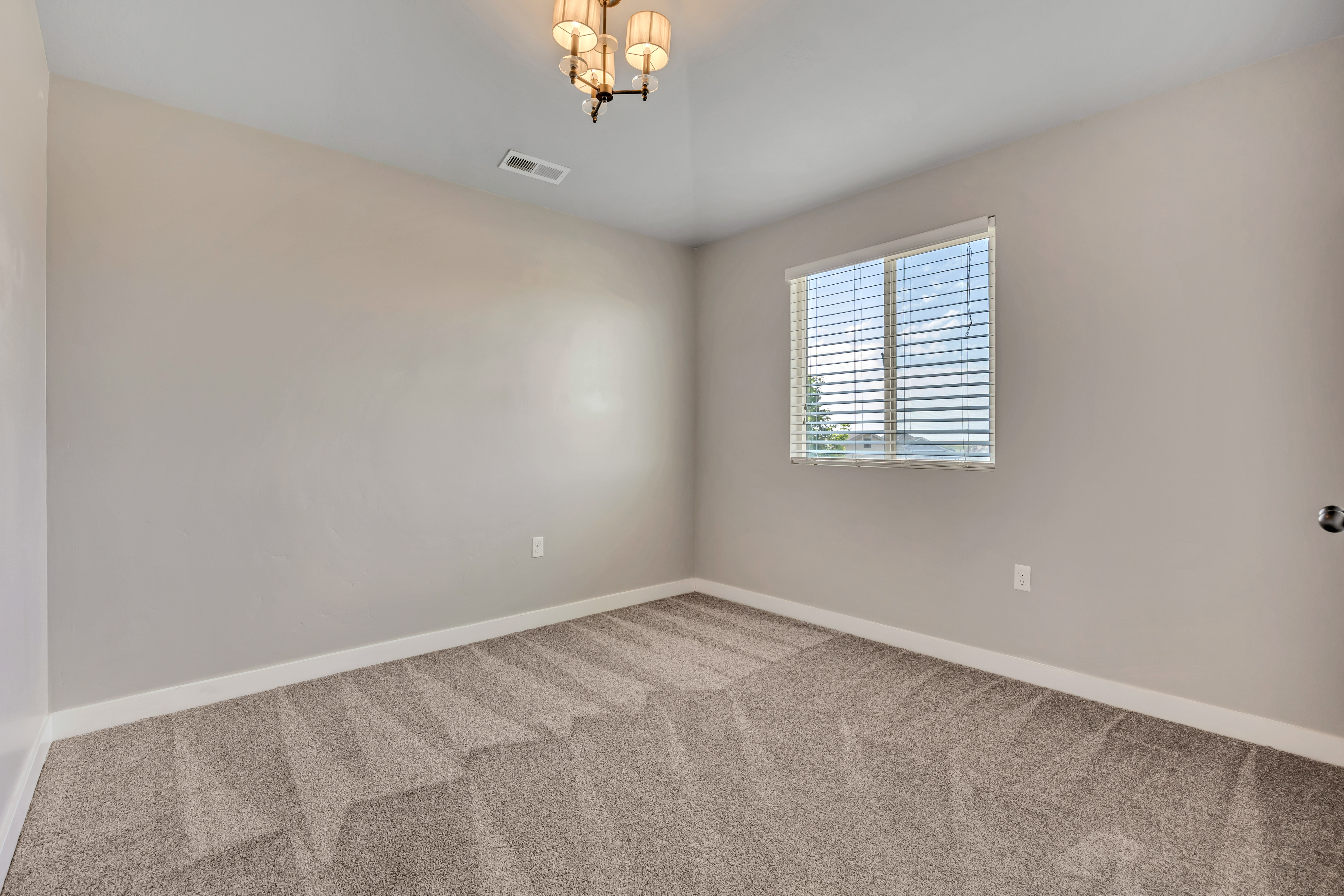 Spacious room featuring neutral wall paint, light-colored patterned carpeting, a single window with blinds, and a decorative ceiling light fixture