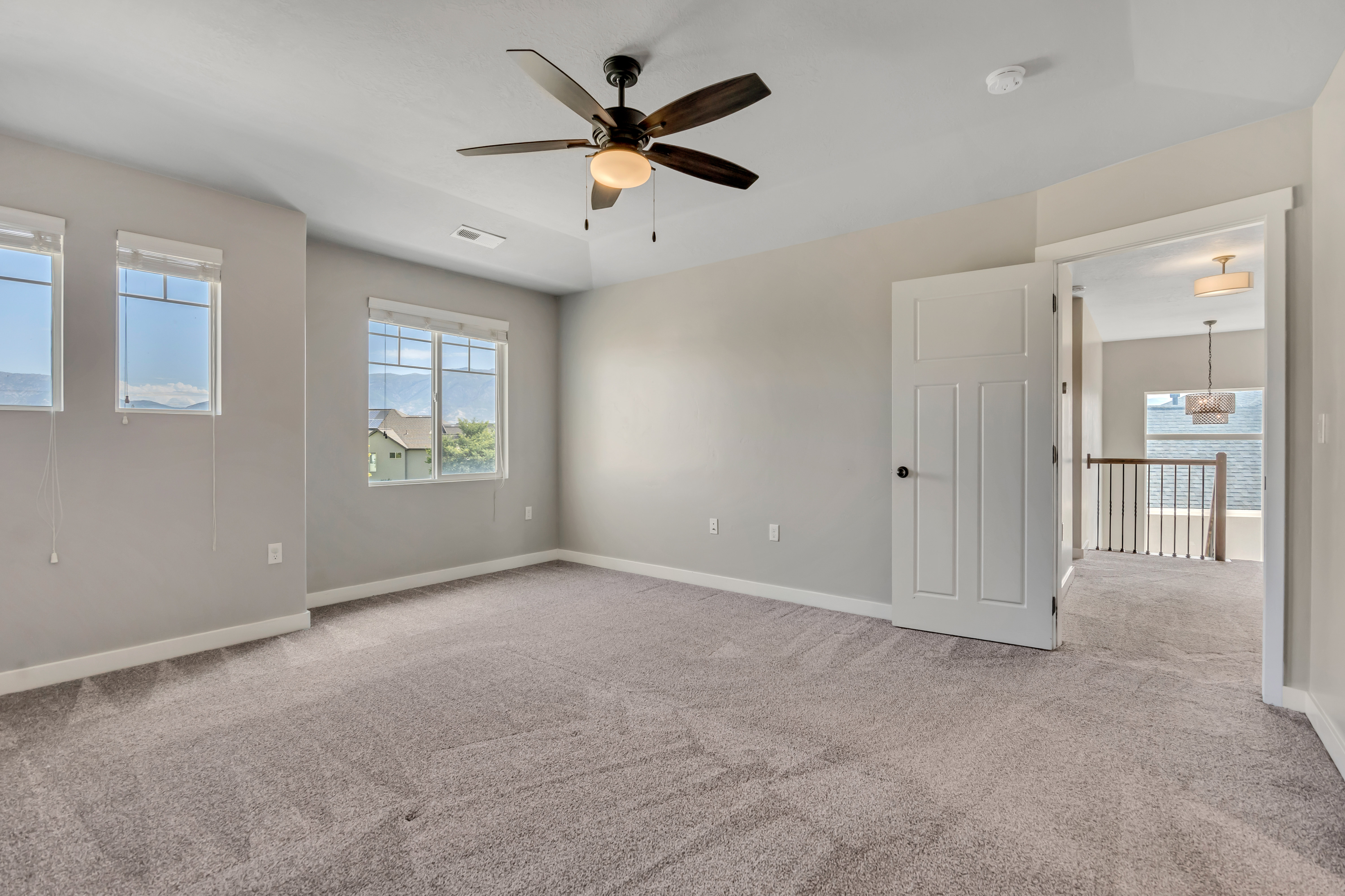 Spacious carpeted room featuring a ceiling fan with integrated lighting, multiple windows, and a solid core door with matte black hardware