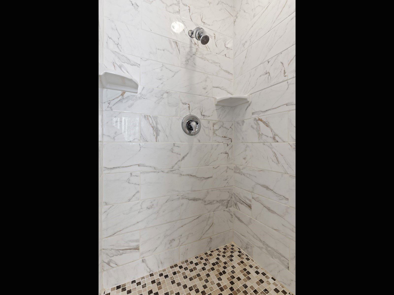 Shower stall featuring white marble-look subway tile walls with subtle gray veining, a mosaic tile floor in neutral tones, a built-in shower niche, and a chrome shower head and faucet