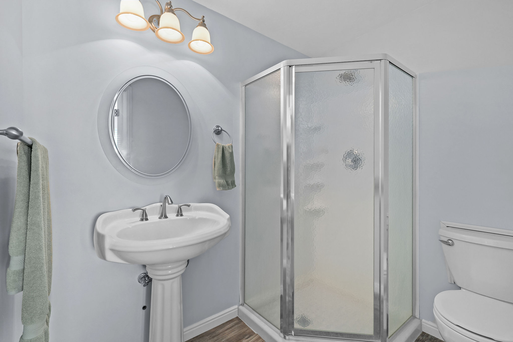 Compact bathroom featuring a pedestal sink with brushed nickel fixtures, a round wall mirror, and a three-light vanity fixture