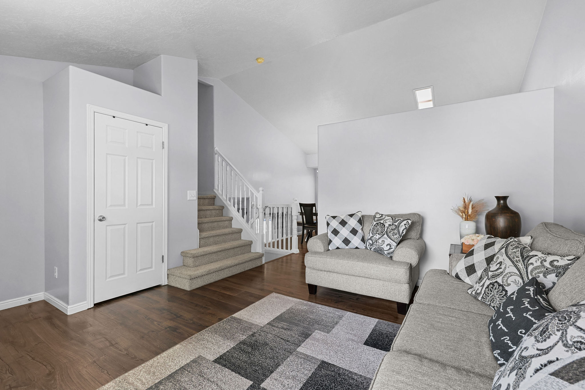 Vaulted ceiling living area featuring wood-finish flooring, a carpeted staircase with white railings, and a six-panel interior door