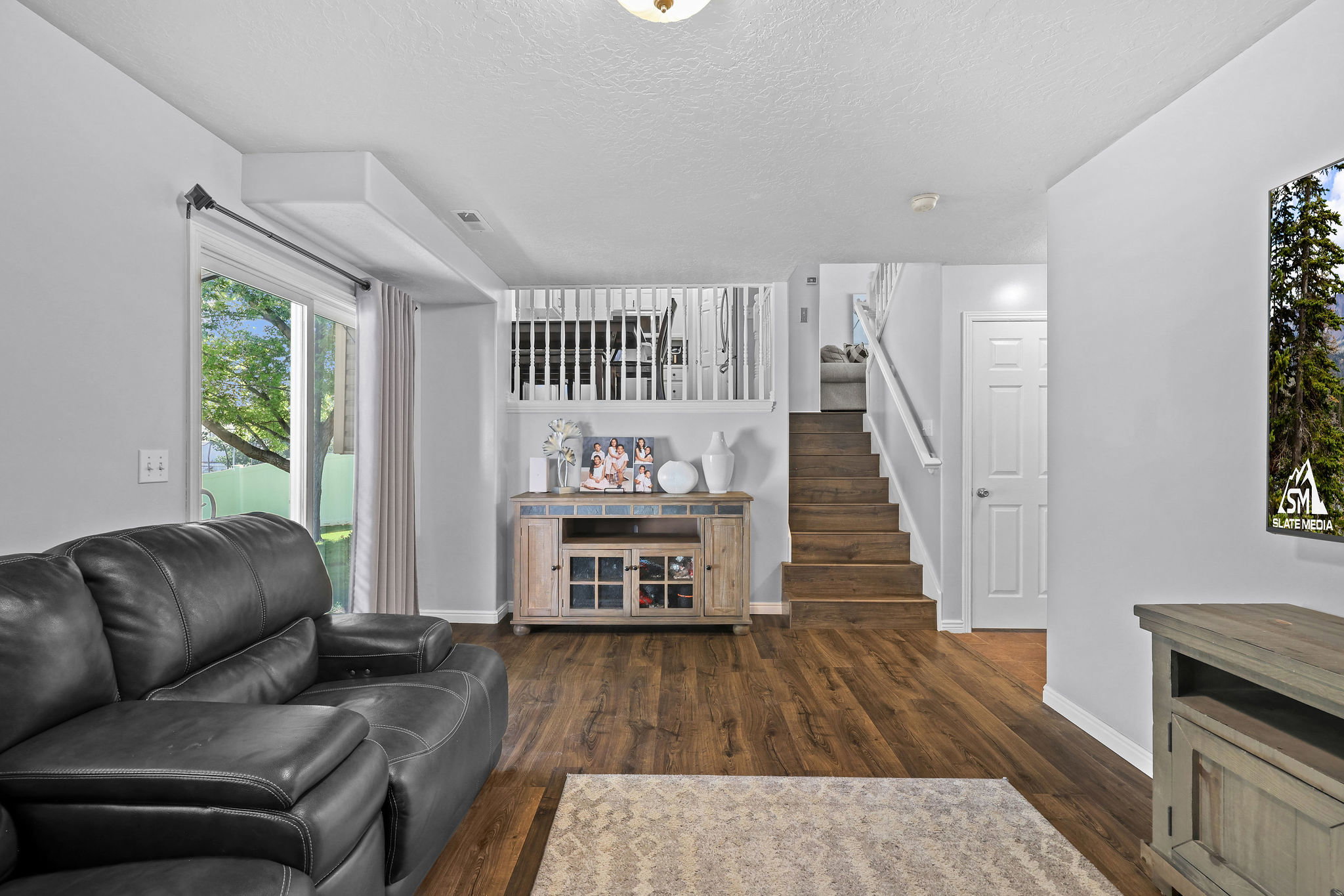 Spacious living area featuring rich wood-finish flooring, a sliding glass door providing outdoor access, and a contemporary white staircase with matching railing
