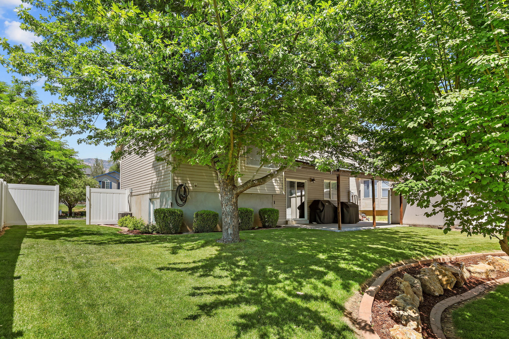 Expansive fenced backyard featuring a substantial shade tree, a rock-lined garden bed, and a concrete patio
