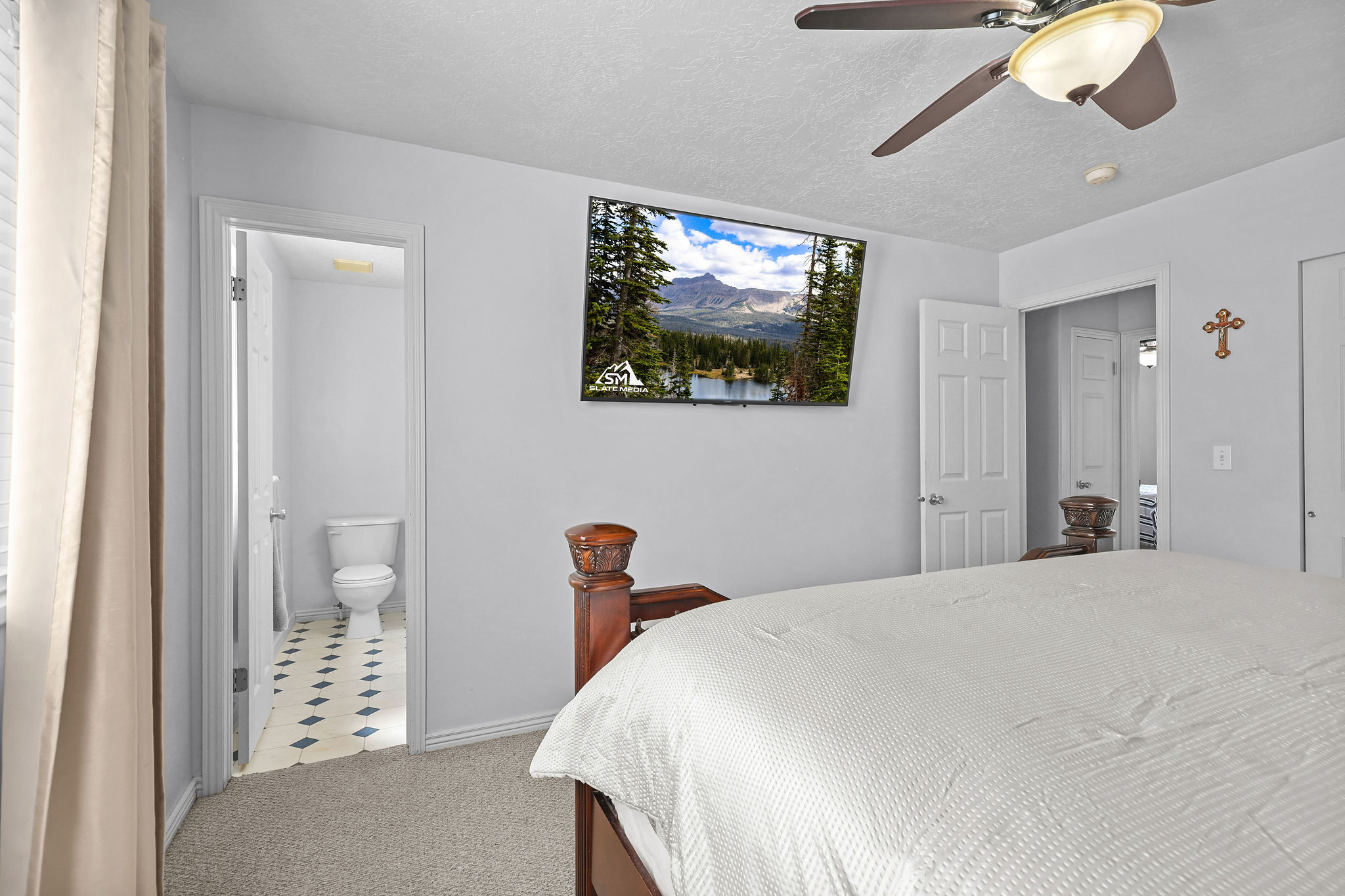 Carpeted bedroom featuring a ceiling fan with integrated lighting, a wall-mounted television, and a doorway leading to a bathroom with patterned floor tiles and a toilet