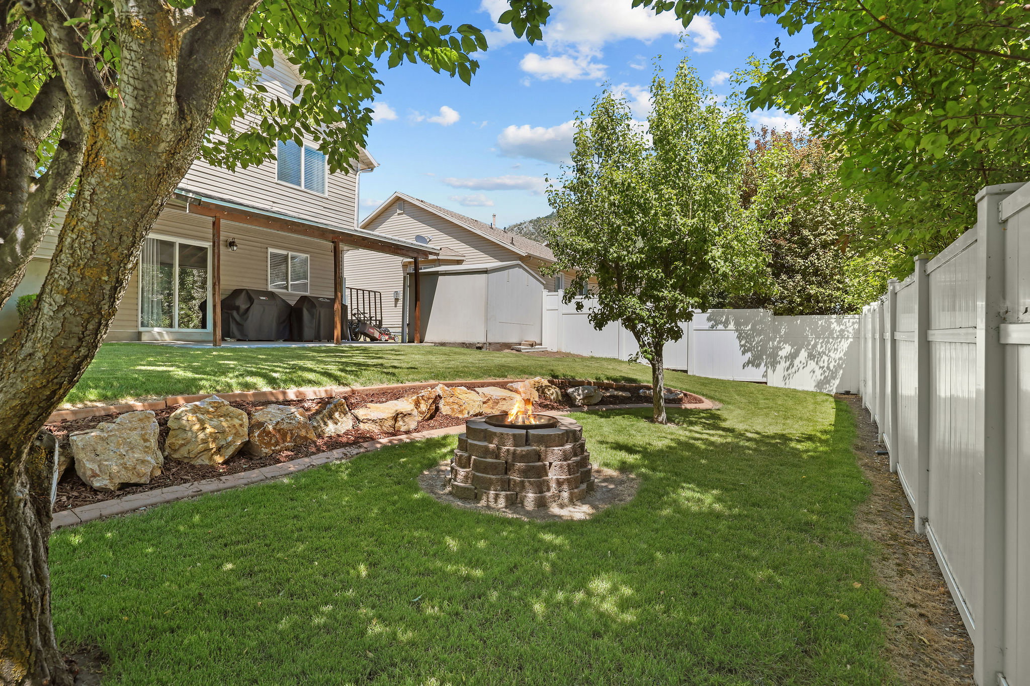 Manicured lawn featuring a brick fire pit, a rock-lined garden bed, a covered patio, and a white privacy fence
