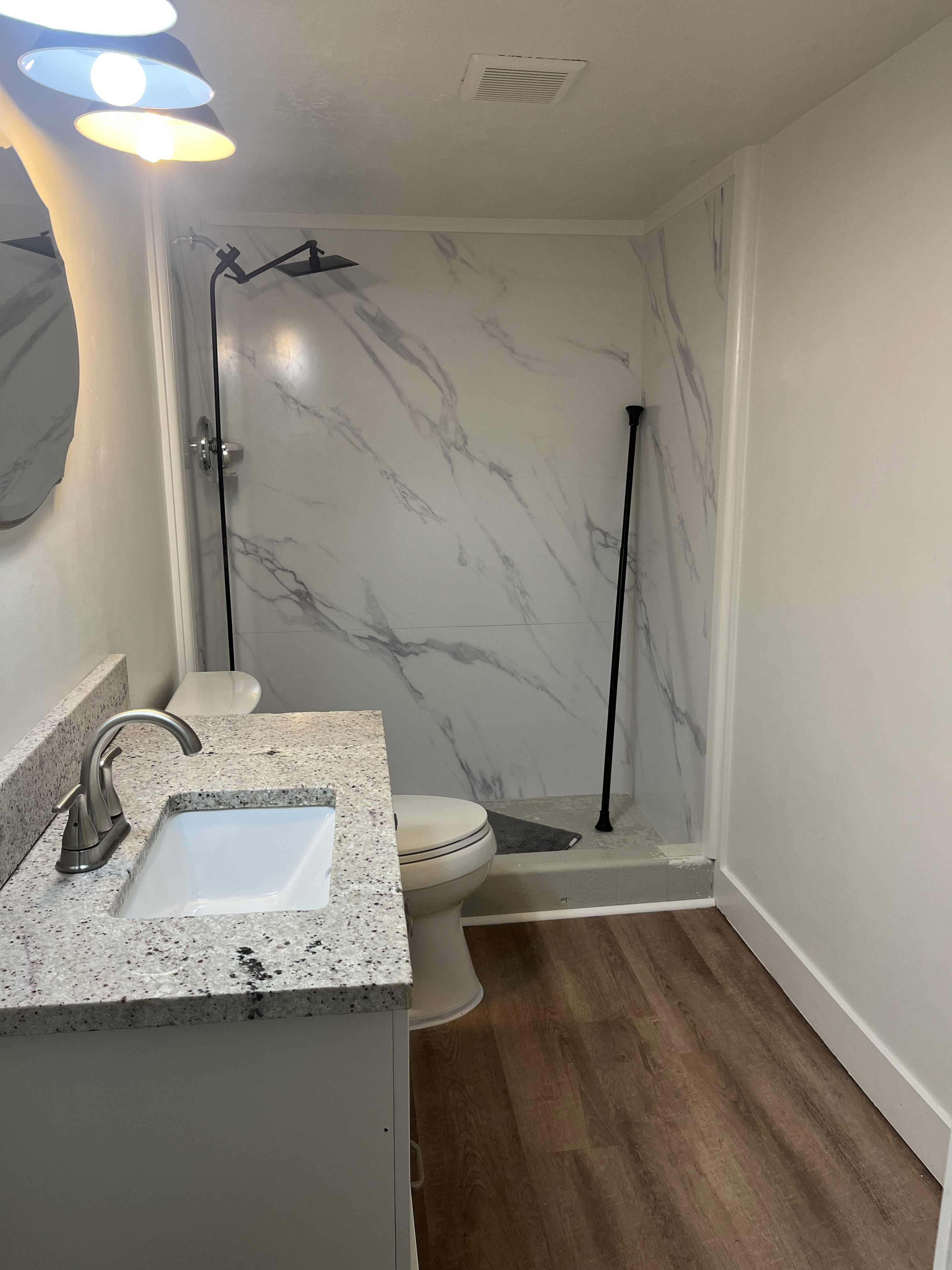 Bathroom featuring a modern vanity with a speckled countertop and undermount sink, wood-finish flooring, a white toilet, and a stand-up shower with marble-look paneling and a matte black shower system