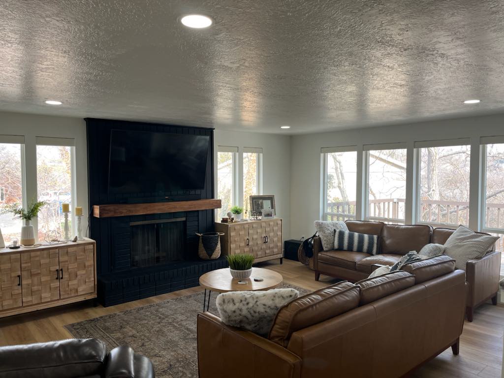 Spacious living area featuring a dark brick fireplace with a wood mantle, numerous large windows, recessed ceiling lighting, and wood-finish flooring