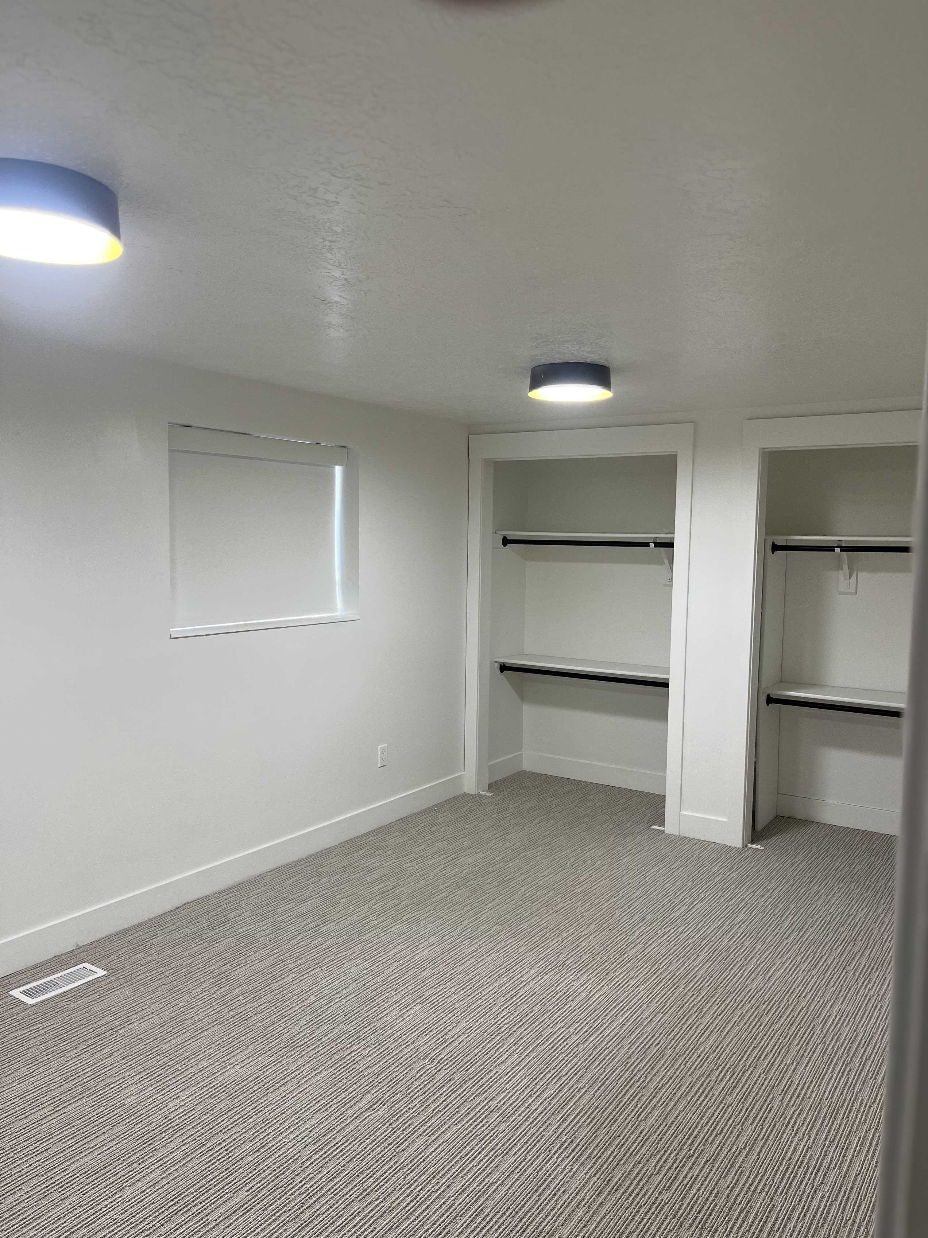 Room featuring two open closets with shelving and hanging rods, patterned carpet flooring, a window with a roller shade, and two flush-mount ceiling lights