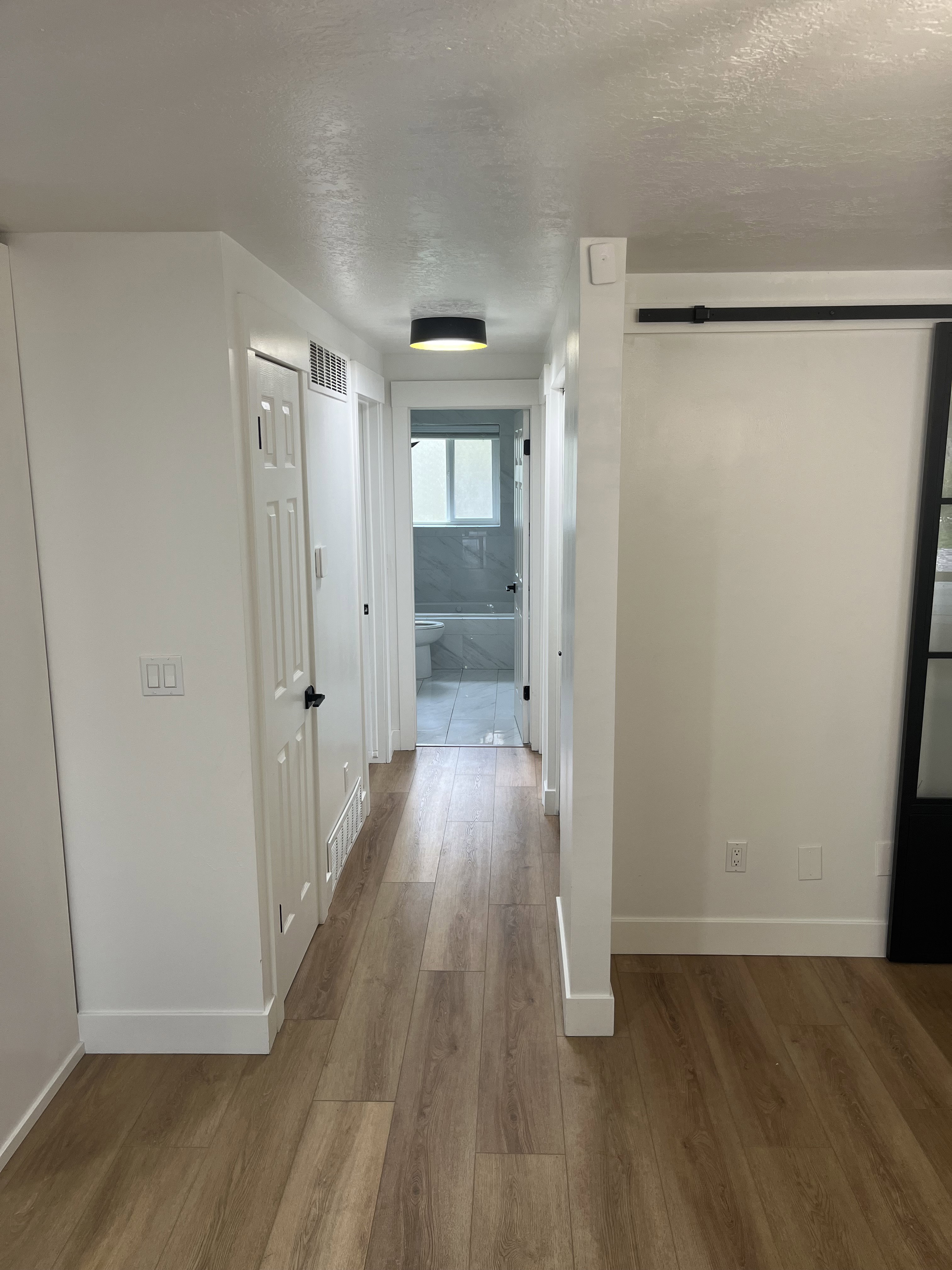 Hallway featuring wood-finish flooring, white baseboards, a black ceiling light fixture, and a black barn door track