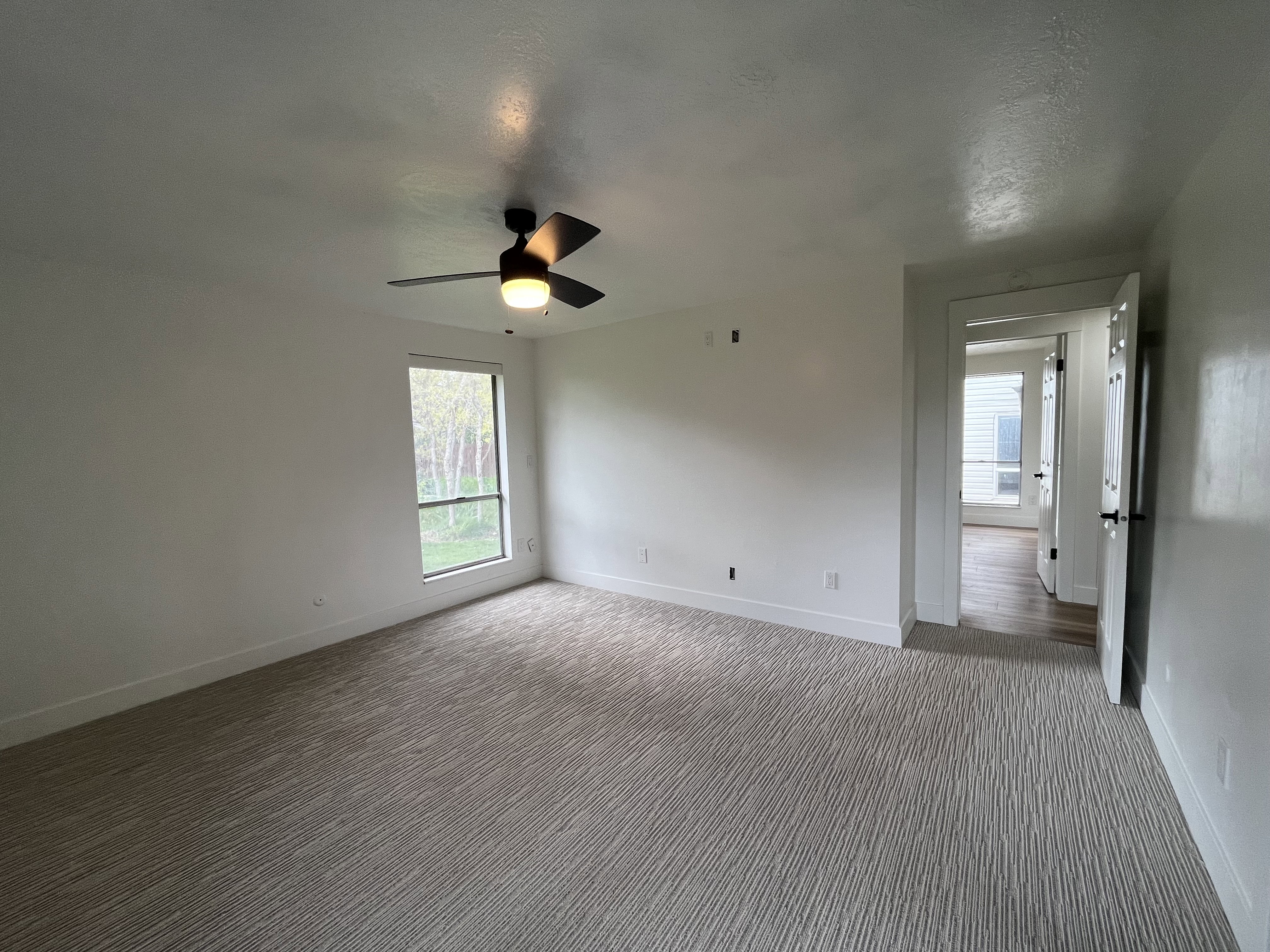 Spacious room featuring new patterned carpet, a modern black ceiling fan with integrated lighting, and a large window overlooking exterior trees