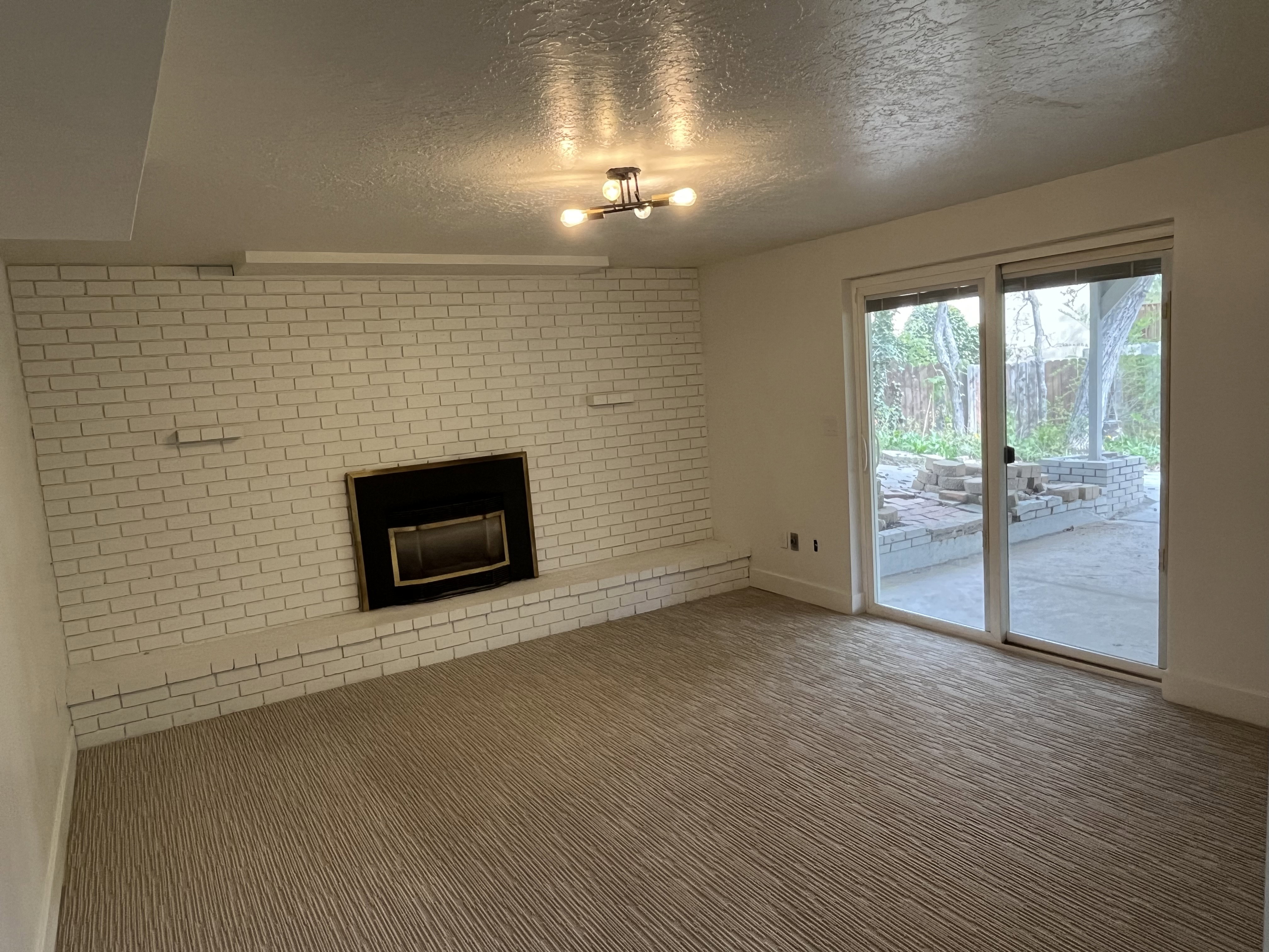 White brick fireplace with a black firebox, light-toned patterned carpeting, and sliding glass doors leading to an outdoor patio with stone hardscaping