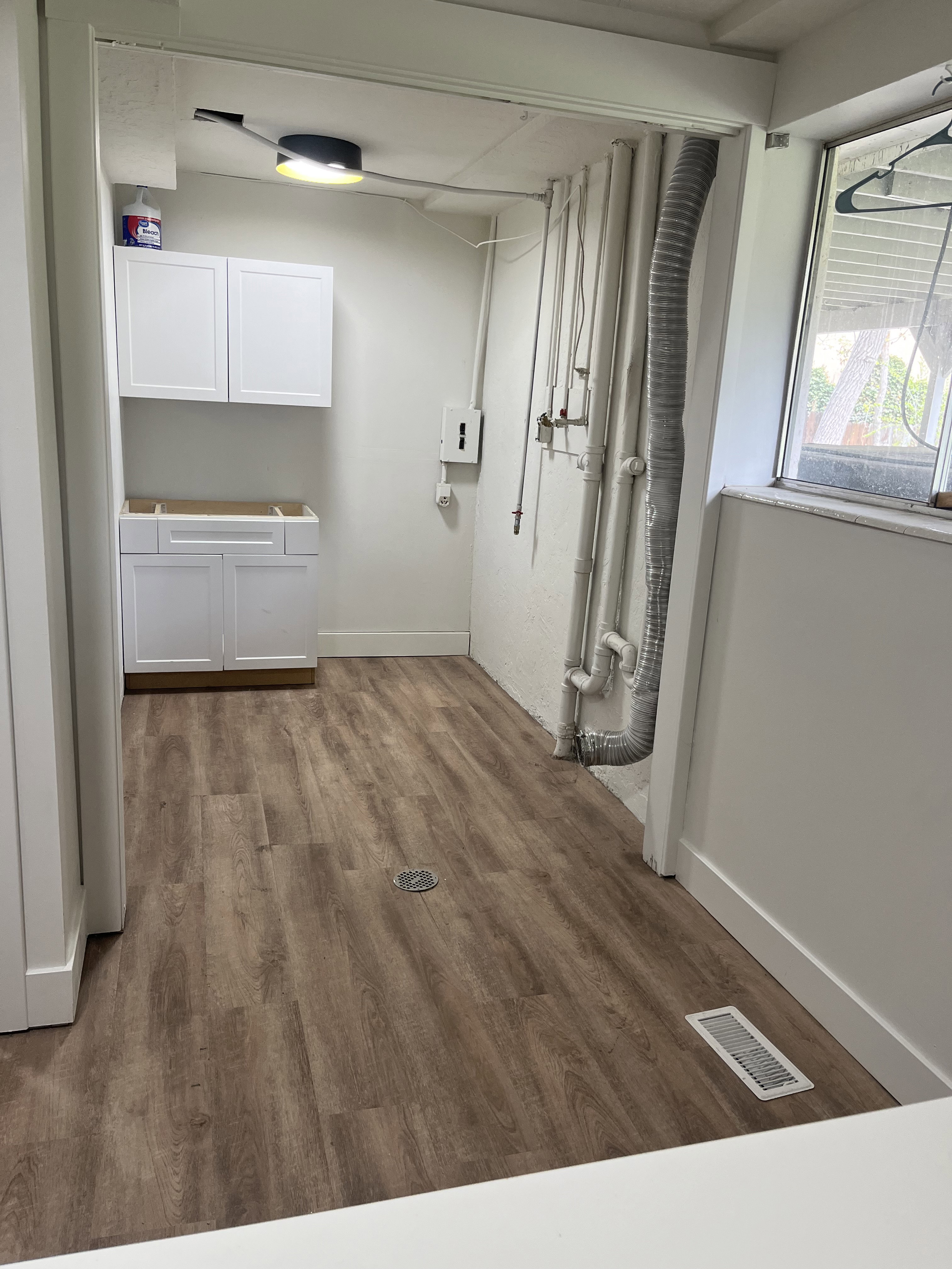 Utility room featuring wood-finish flooring, white cabinetry, overhead lighting, a window, and exposed piping