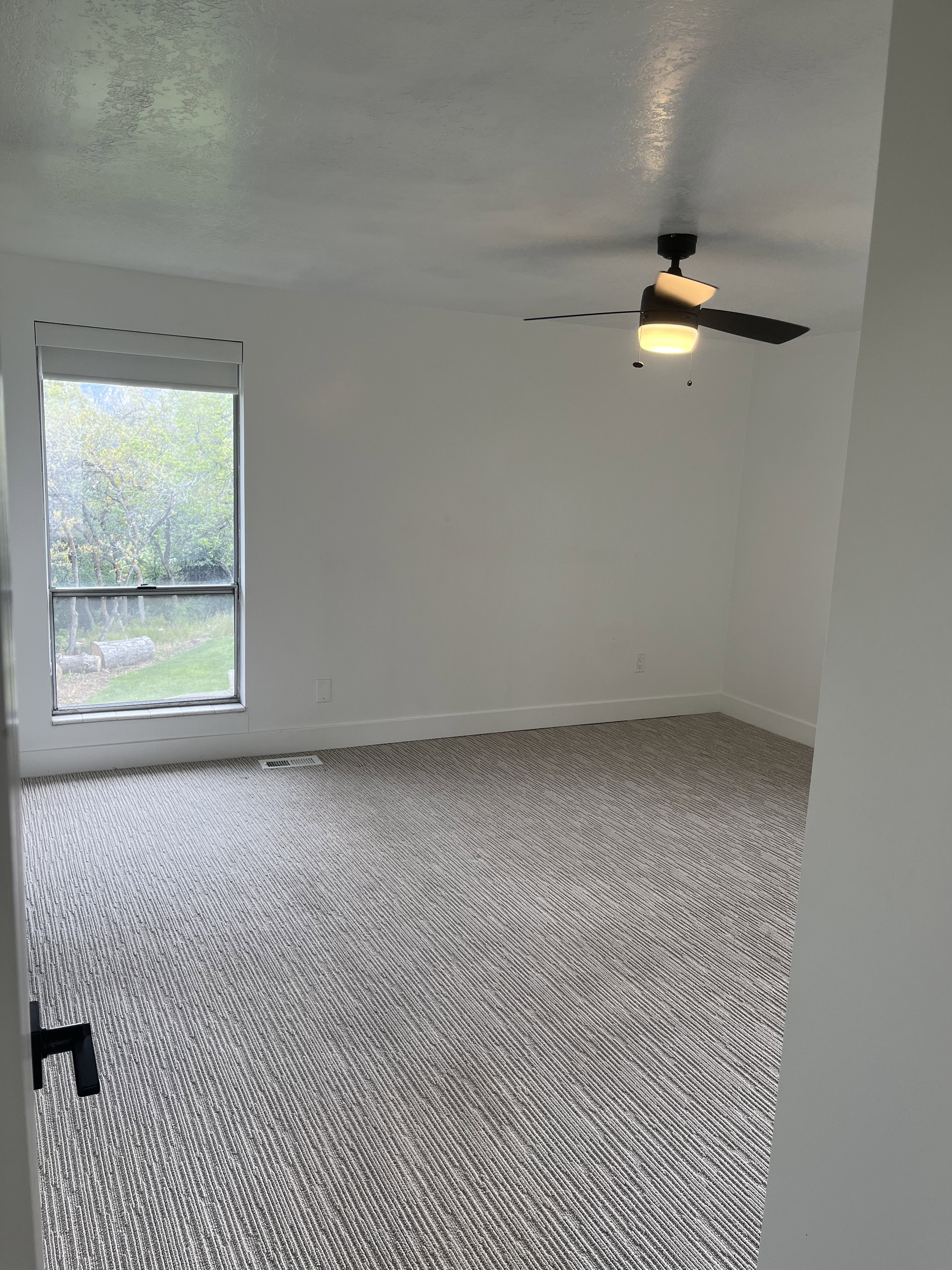 Spacious room featuring neutral carpeting, white walls, a large window, a ceiling fan with integrated lighting, and white baseboards