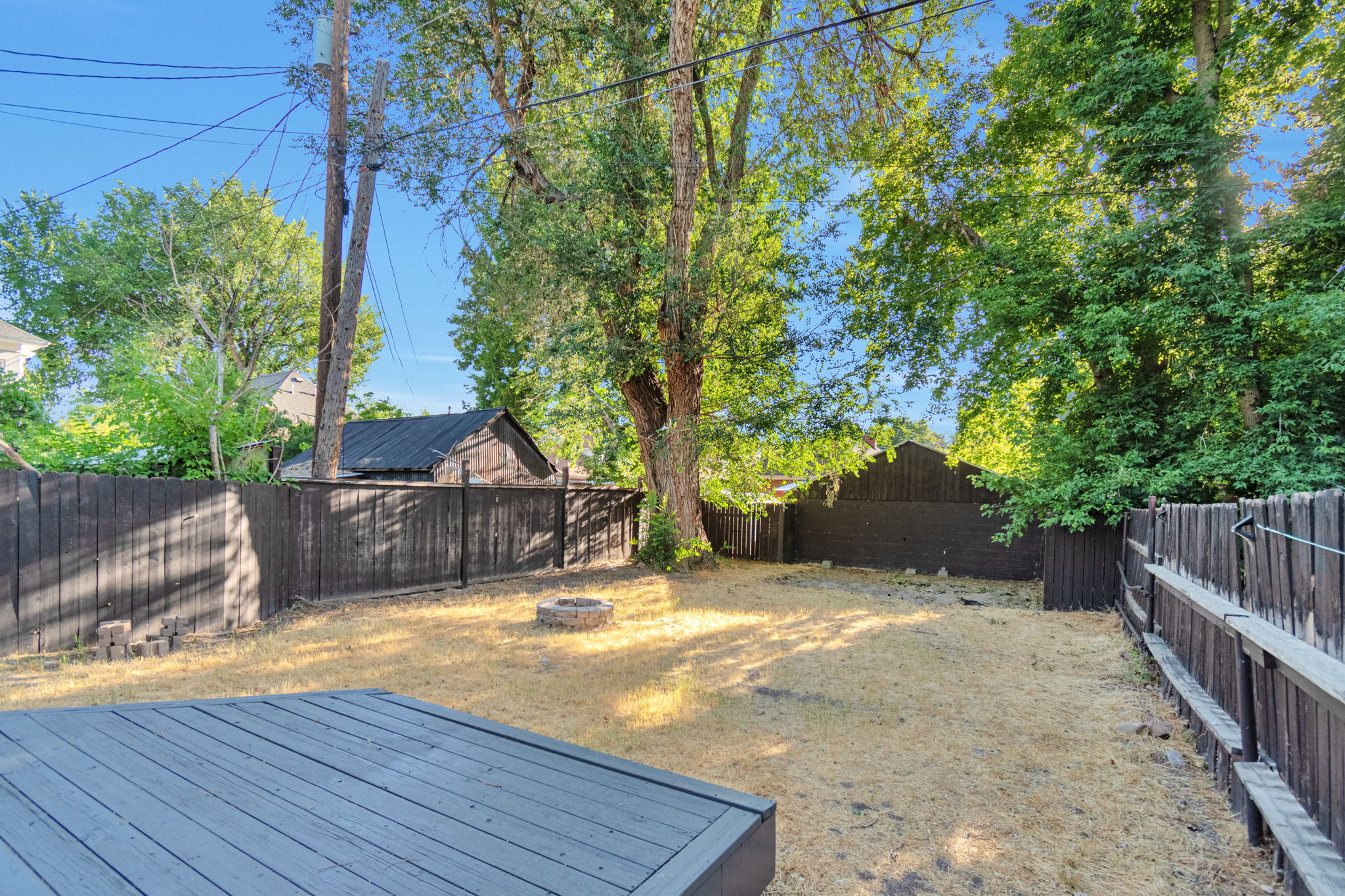 Expansive backyard featuring a gray-painted deck, dark wood fencing, mature trees, and a circular fire pit