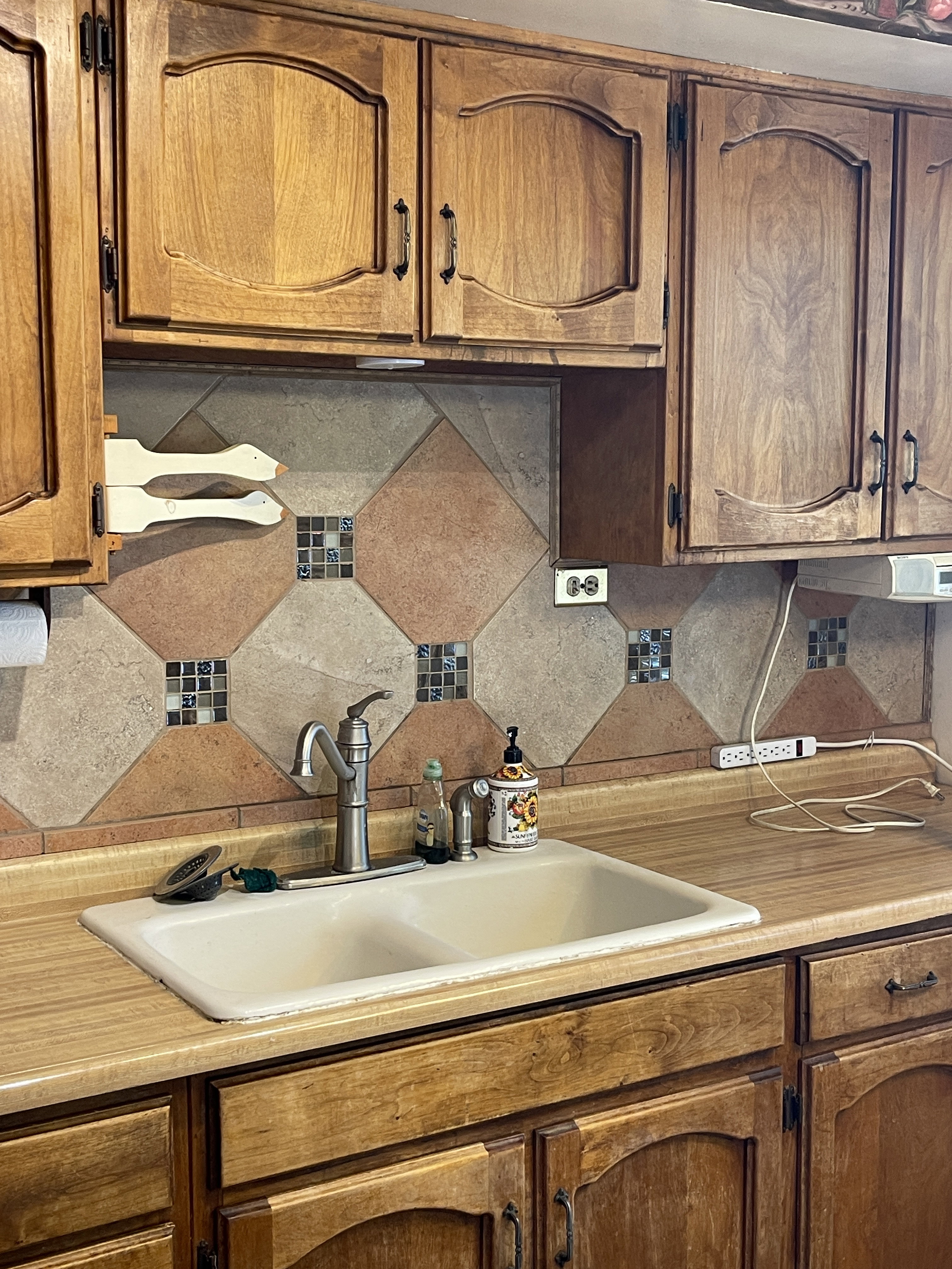Kitchen sink area featuring light wood-finish cabinetry, a neutral-toned countertop, and a tiled backsplash with decorative accents