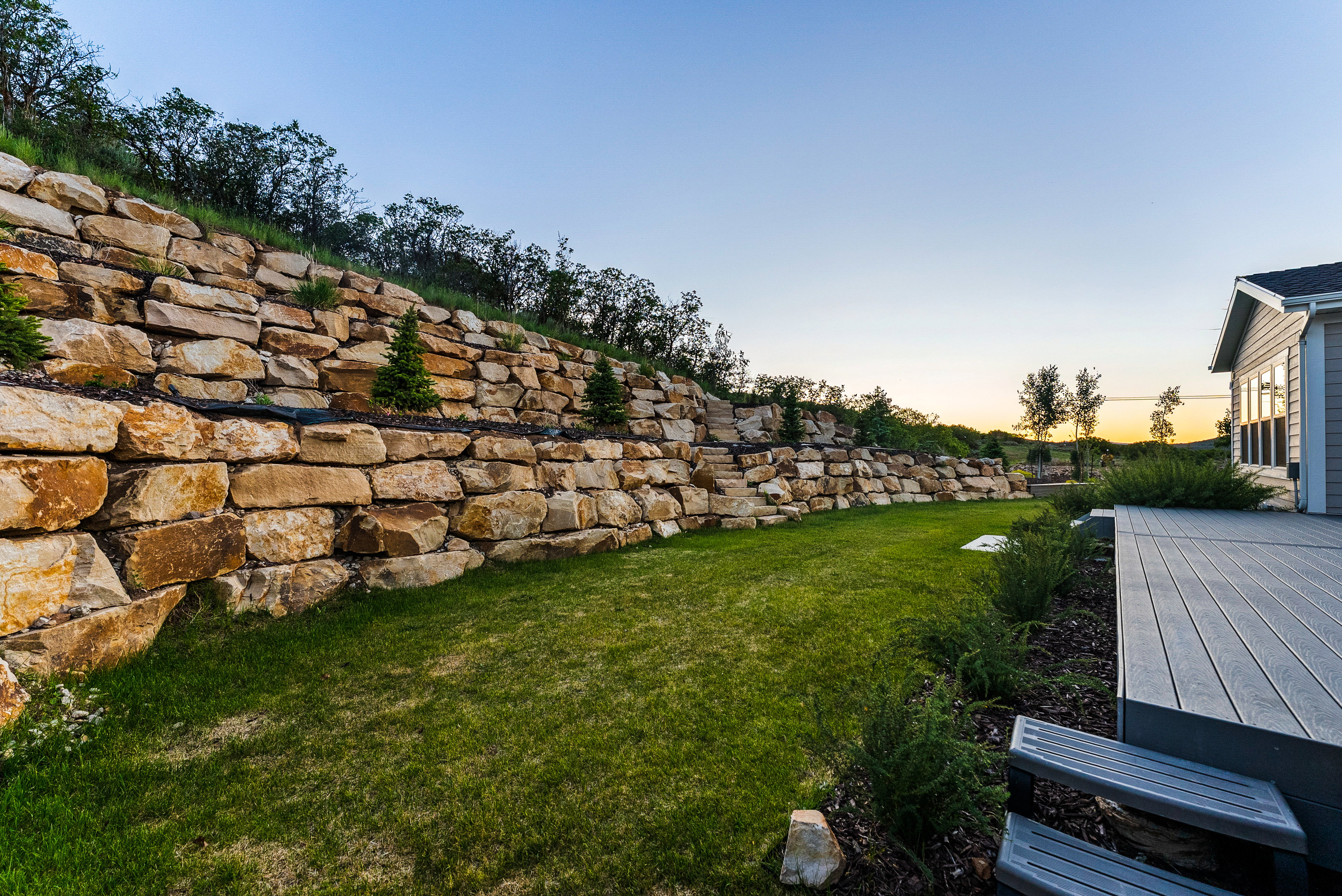 Outdoor area featuring a stone retaining wall, green lawn, composite decking, and mature landscaping