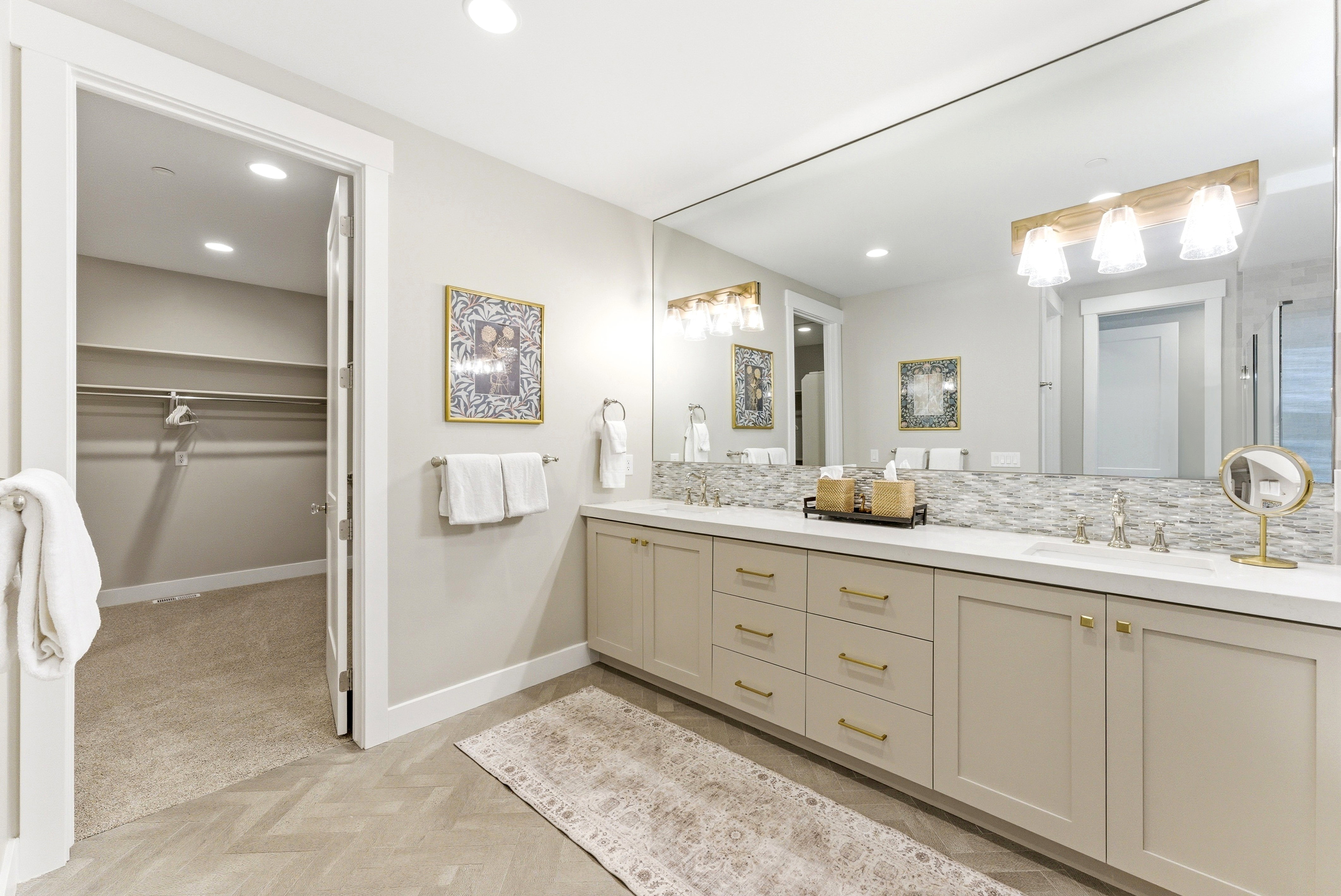 Dual vanity with light cabinetry, gold-toned hardware, solid surface countertops, and a full-width mirror