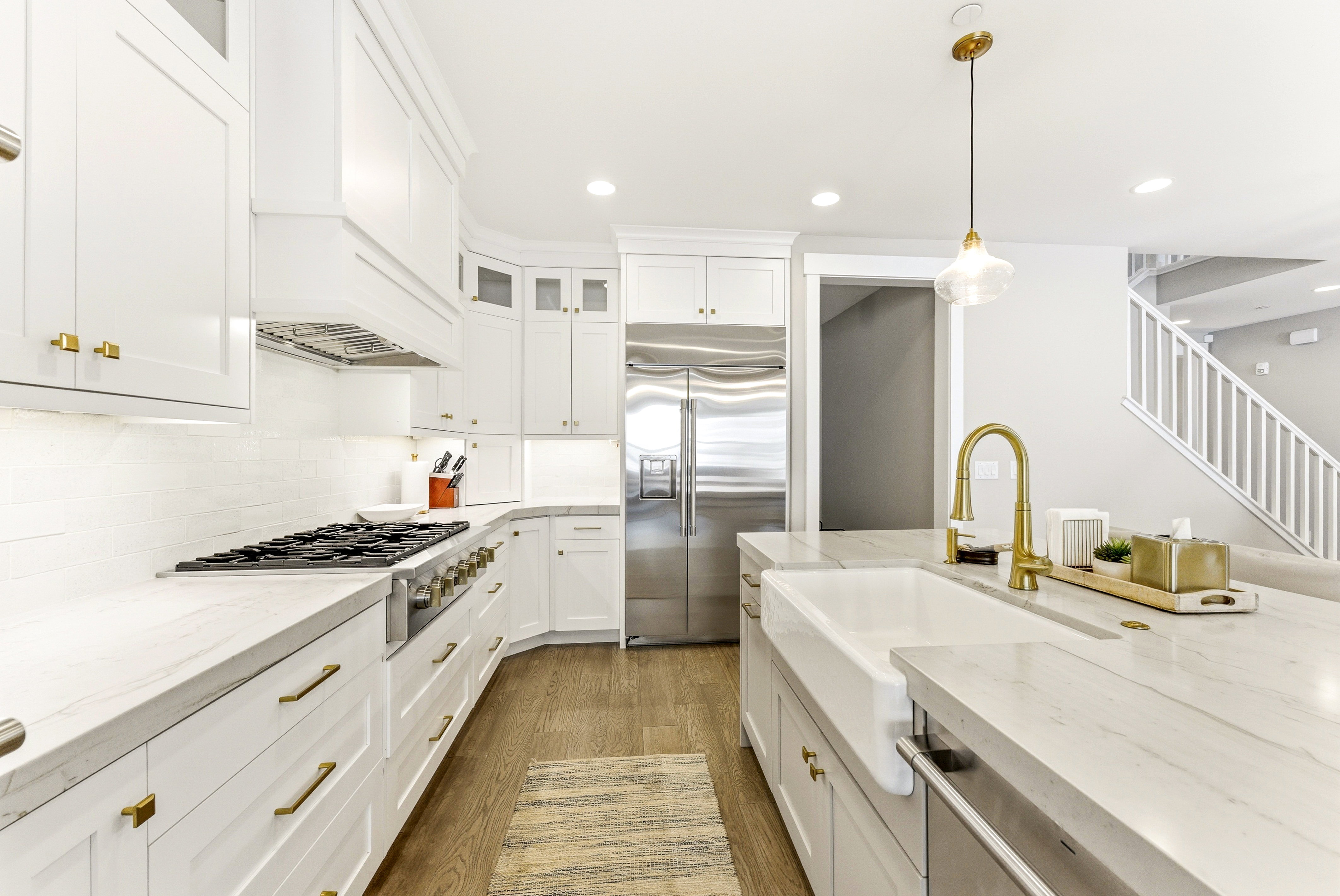 Bright kitchen featuring white cabinetry with brass hardware, a stainless steel refrigerator, a gas range with a matching vent hood, and a farmhouse sink with a gold faucet