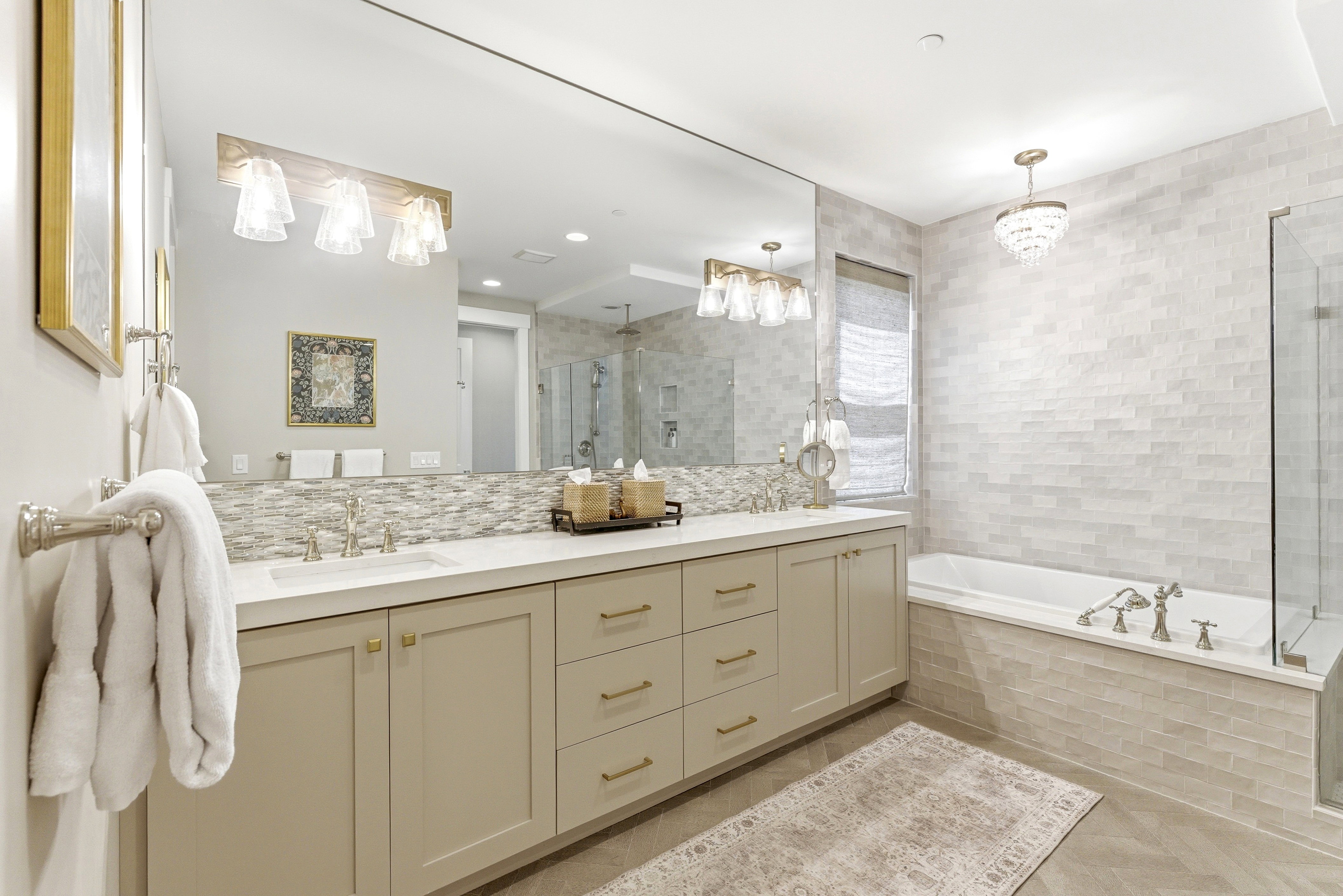 Bathroom featuring an extensive vanity with dual sinks, light-toned cabinetry with brass-finish hardware, a built-in soaking tub, a frameless glass shower enclosure, and light-toned tile work throughout