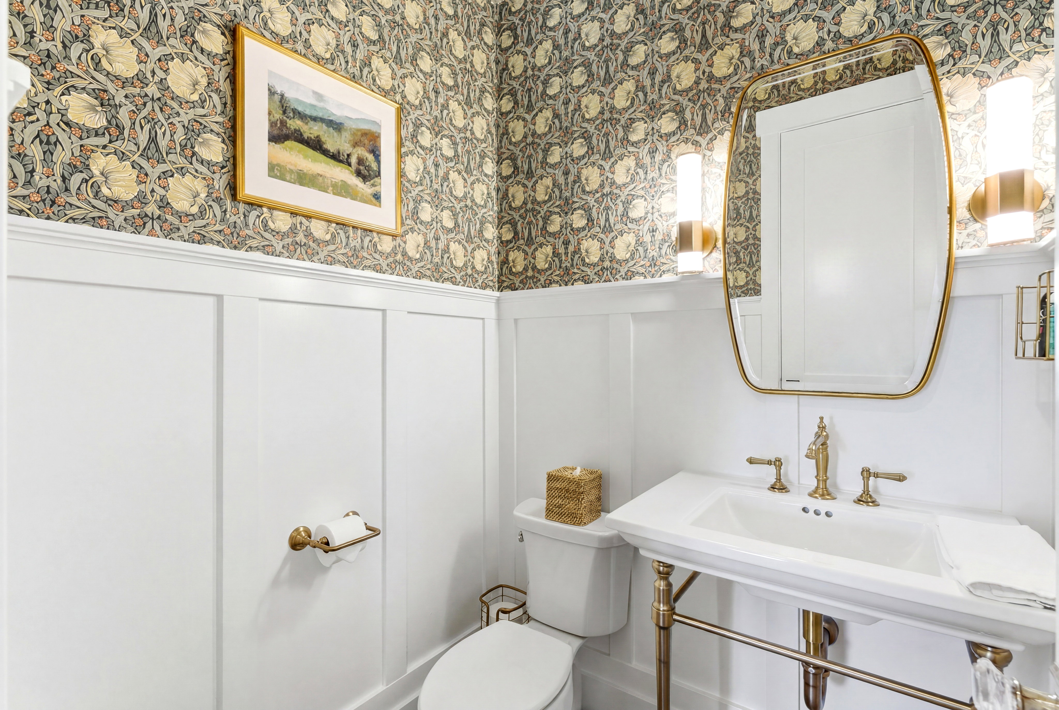Powder room featuring board and batten wainscoting, botanical pattern wallpaper, and a pedestal sink with brass-finish fixtures