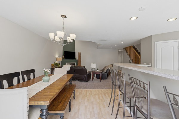 Spacious interior featuring a wood-finish dining table with chandelier, a breakfast bar with light-toned countertop, recessed lighting, and a visible staircase with wooden balusters