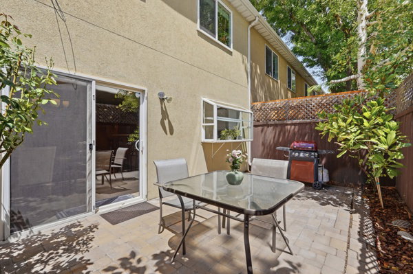 Paved patio area featuring light-colored pavers, surrounded by mature greenery and a wood privacy fence