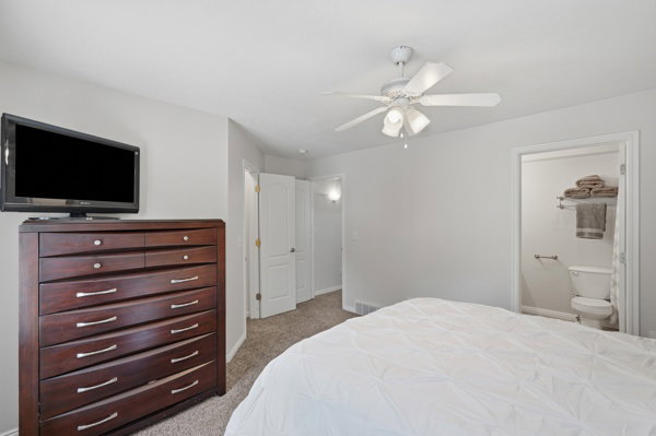 Bedroom featuring light grey walls, carpet flooring, a ceiling fan with integrated lighting, and an open doorway revealing a partial bathroom with a toilet and grab bar