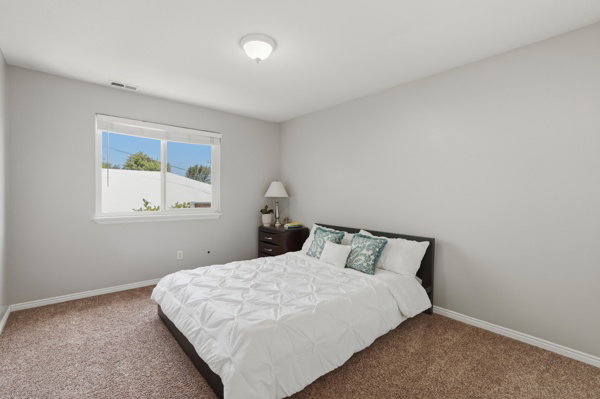 Bright bedroom with a large window, neutral gray walls, brown carpet flooring, white baseboards, and a flush-mount ceiling light fixture