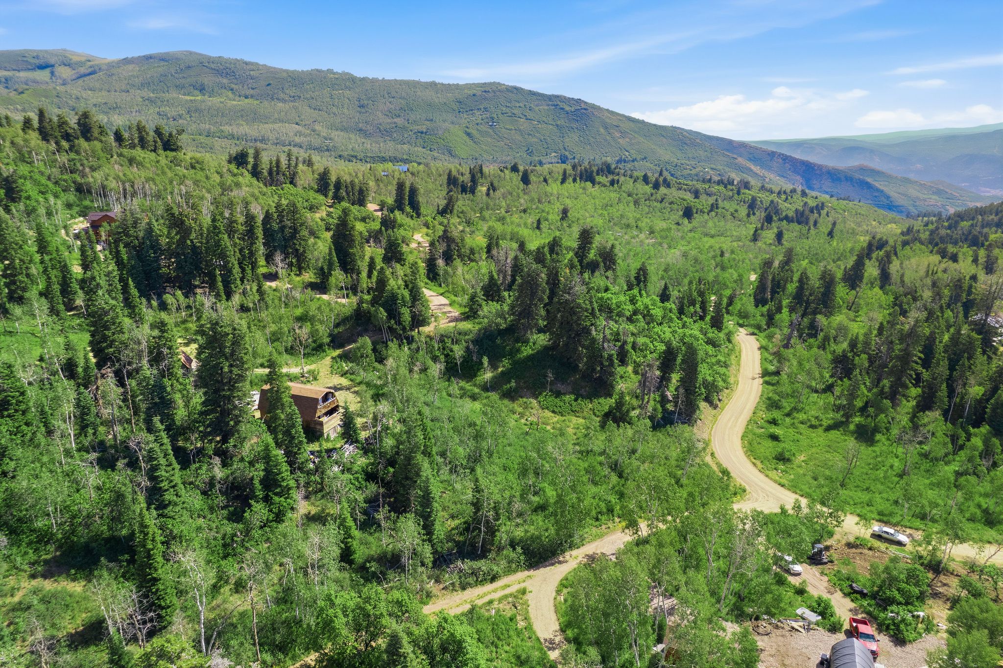 Expansive mountain landscape featuring dense evergreen and deciduous trees, a winding unpaved road, and distant mountain ranges