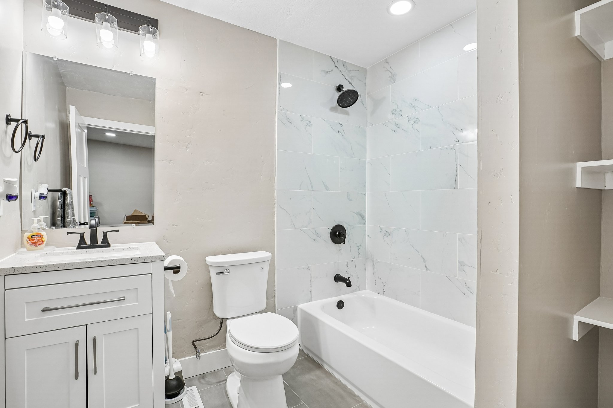 Bathroom featuring a white vanity with a speckled countertop, a modern white toilet, and a bathtub with a white marble-look tiled surround