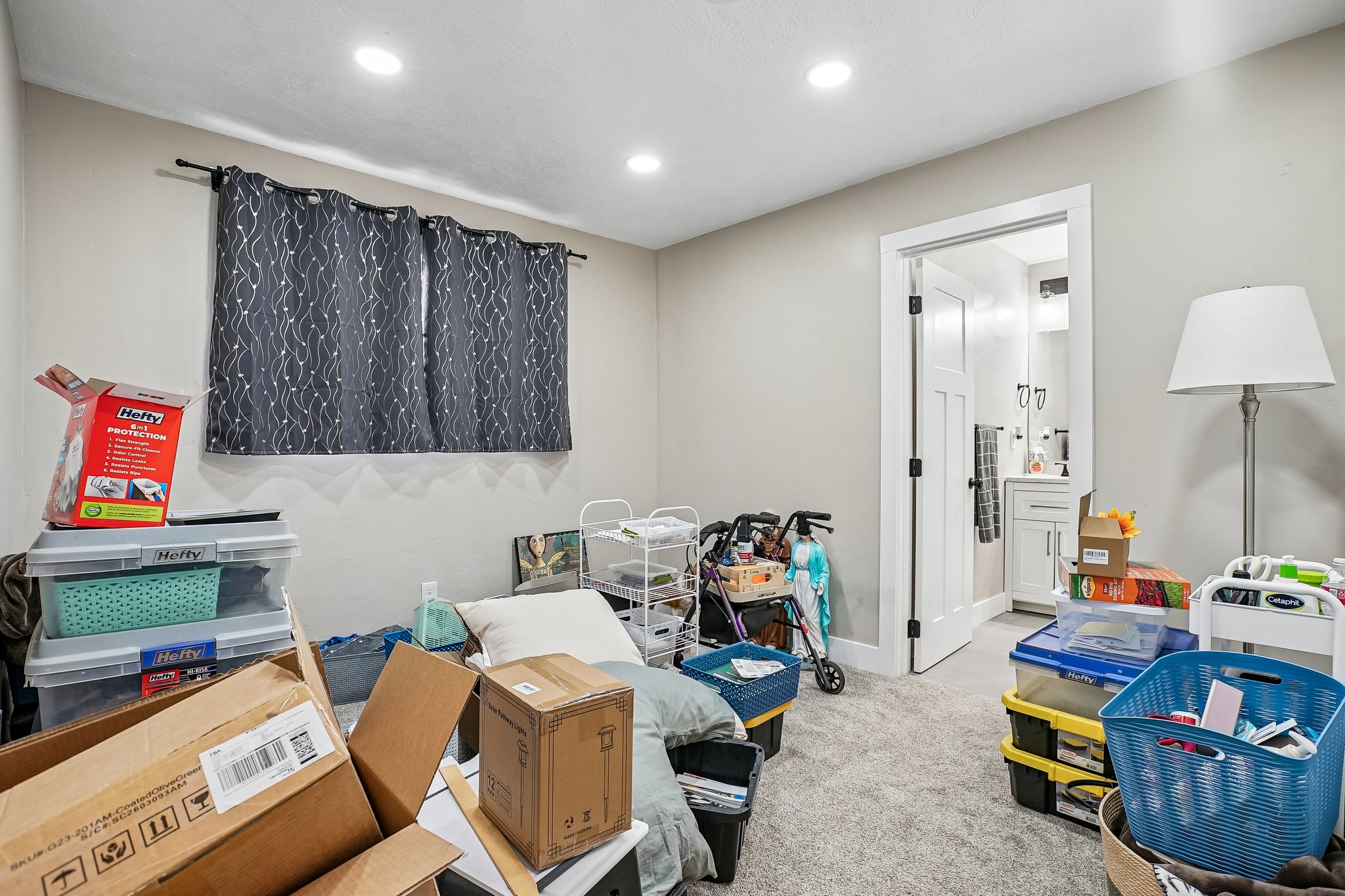Recessed lighting illuminates a neutral-toned room featuring a window with dark patterned curtains and light gray walls