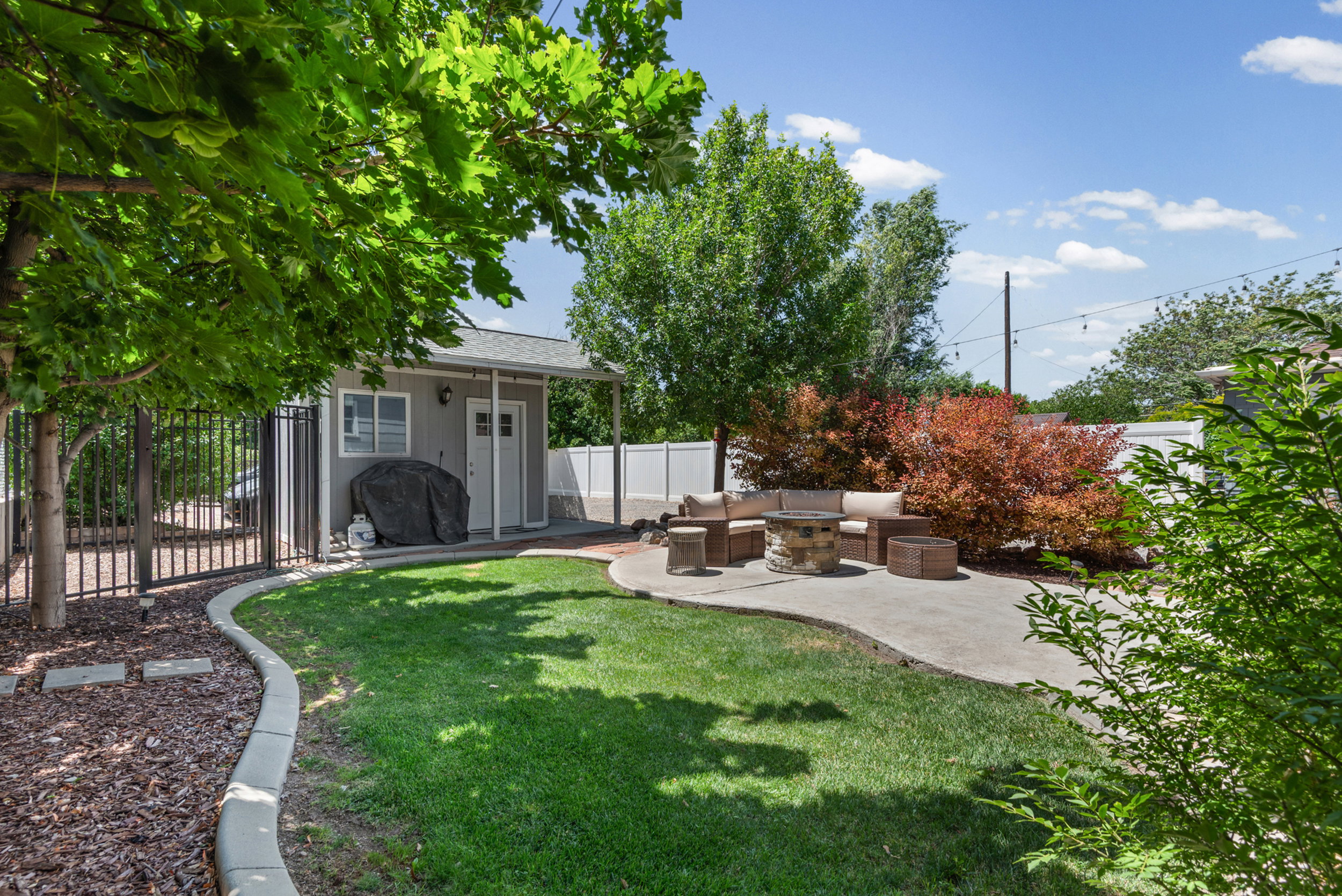 Backyard featuring a concrete patio, grassy lawn with defined edging, mature trees providing shade, and a detached structure with a pitched roof