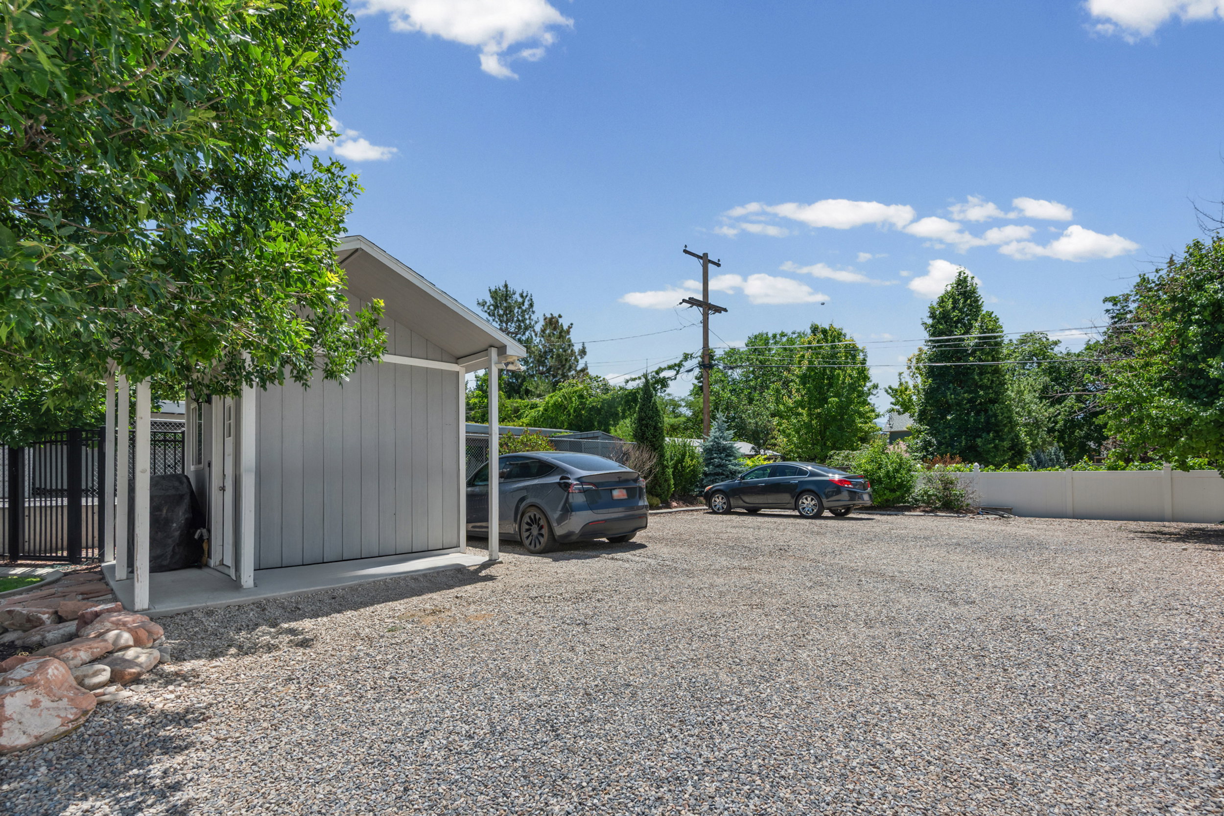 Gravel parking area featuring a covered structure with vertical siding, a solid fence, mature trees, and a distant privacy fence