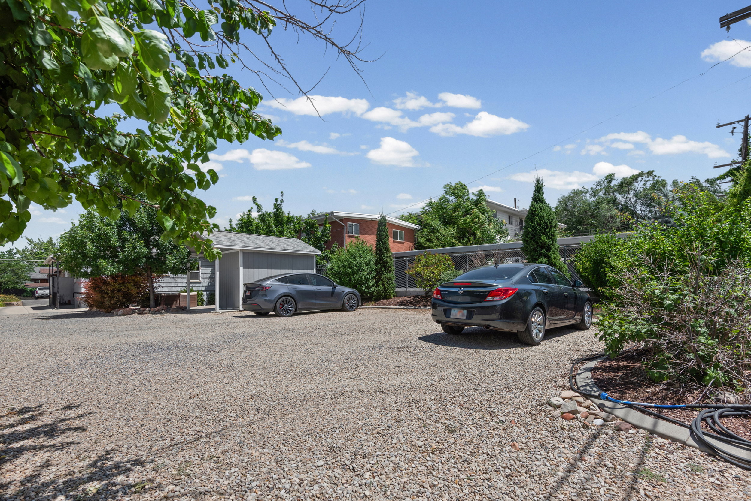 Expansive gravel parking area featuring a detached garage with a light gray facade, a fenced perimeter, and mature trees providing natural shading