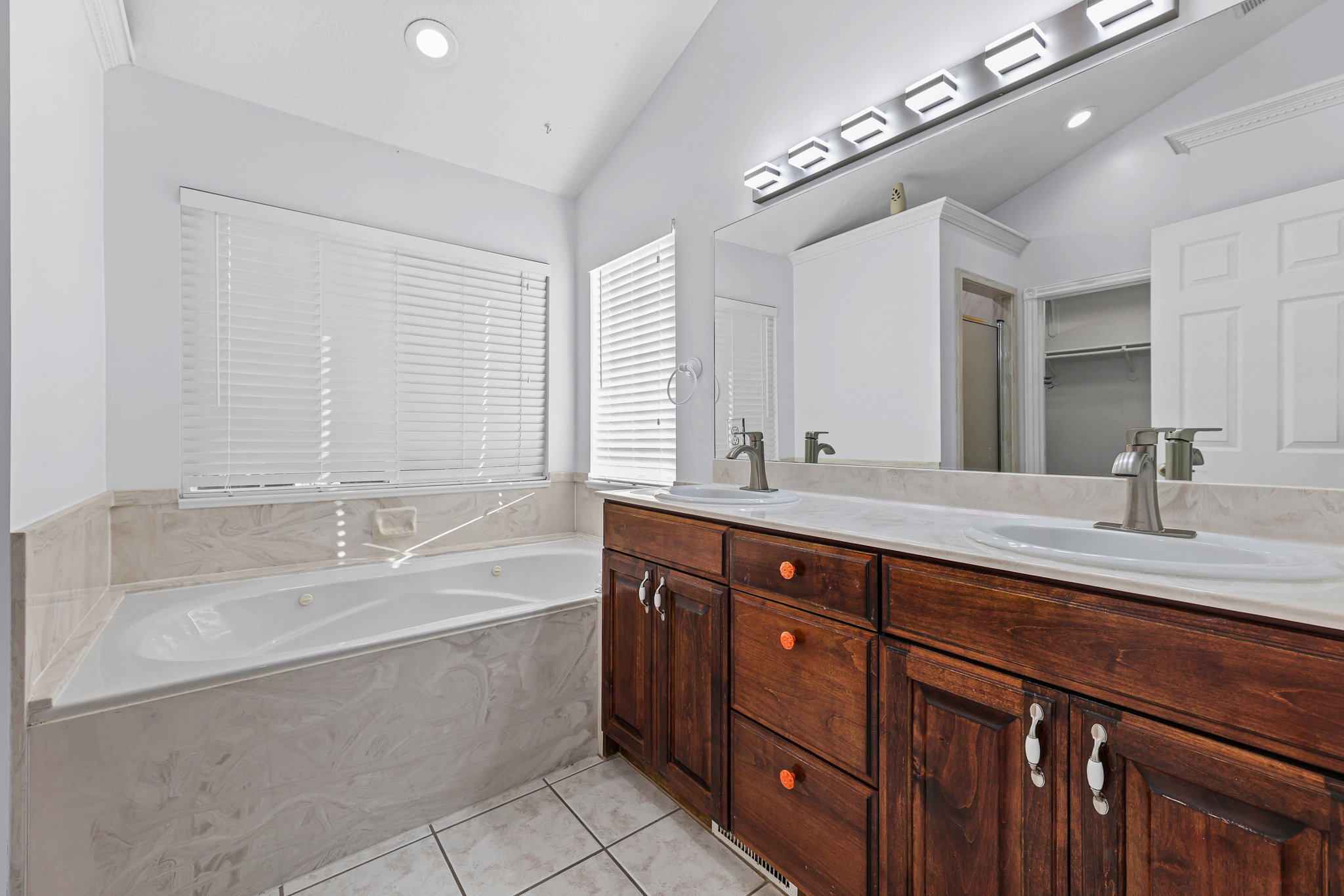 Bathroom featuring a jetted soaking tub with a tiled surround, dual wood-finish vanities, a large mirror, and recessed lighting
