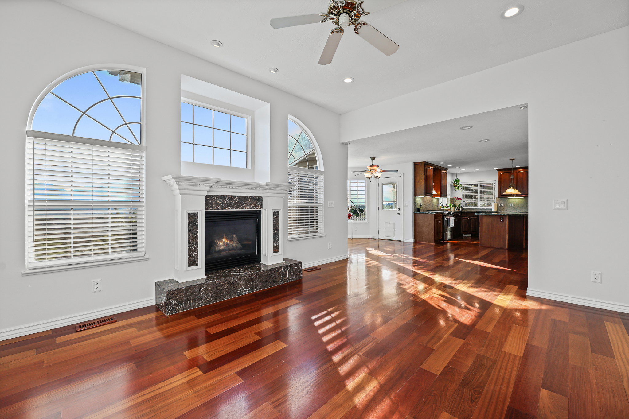 Hardwood flooring throughout a bright living space featuring a fireplace with a stone surround and hearth, framed by arched and rectangular windows