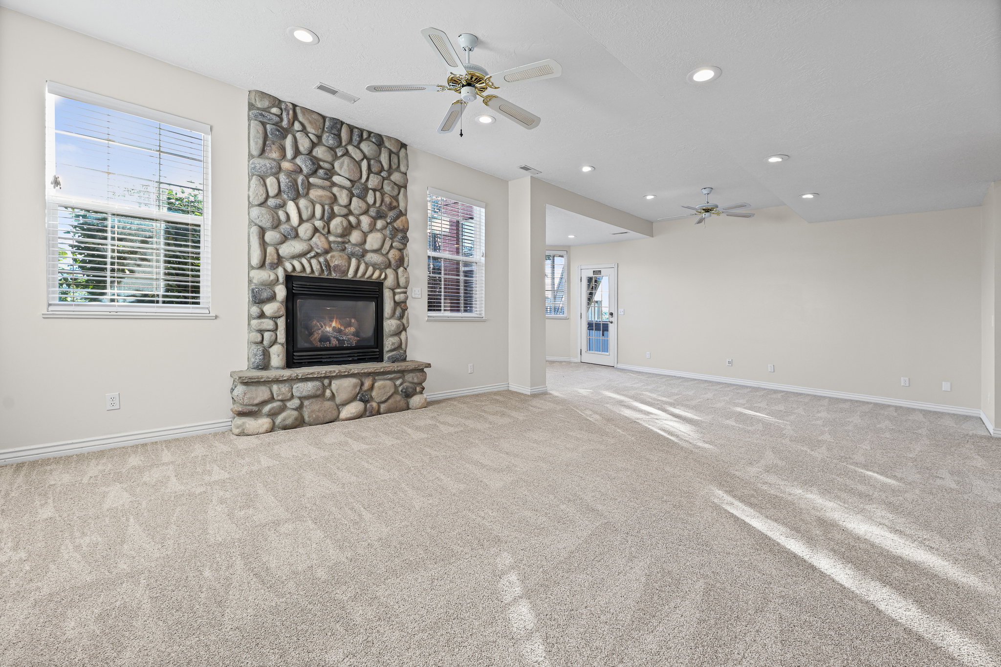 Spacious living area featuring a floor-to-ceiling stone fireplace, plush carpeting, recessed lighting, and multiple windows with blinds